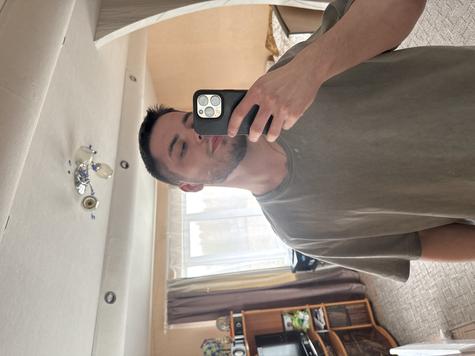 A man with short dark hair and a beard is taking a selfie in a mirror. He is holding a black phone with multiple camera lenses. The reflection shows a room with a window, curtains, a TV, and a bookshelf. The location appears to be in a home, and the city is Бородулино, Russia.
