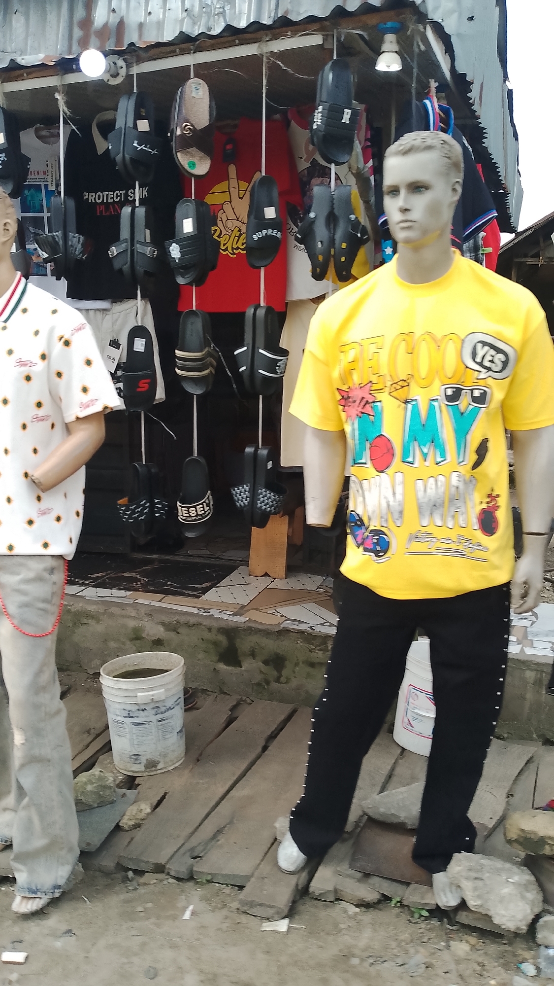 This image depicts an outdoor market stall in Agbarho, Nigeria, likely in the Delta State. The stall features a variety of merchandise, including t-shirts and footwear, displayed on racks and hung from the ceiling. In the foreground, two mannequins are dressed in clothing. The mannequin on the right wears a yellow t-shirt with graphic text and black pants adorned with silver studs. The mannequin on the left is partially visible, wearing a white t-shirt with a sunflower print and grey trousers. Behind the mannequins, numerous pairs of flip-flops and sandals are displayed. A white bucket filled with dark liquid is situated on wooden planks to the left. The overall environment suggests a street-side vendor, with a corrugated metal roof overhead and a rough, possibly unpaved, ground surface. The scene appears to be set during daylight hours.