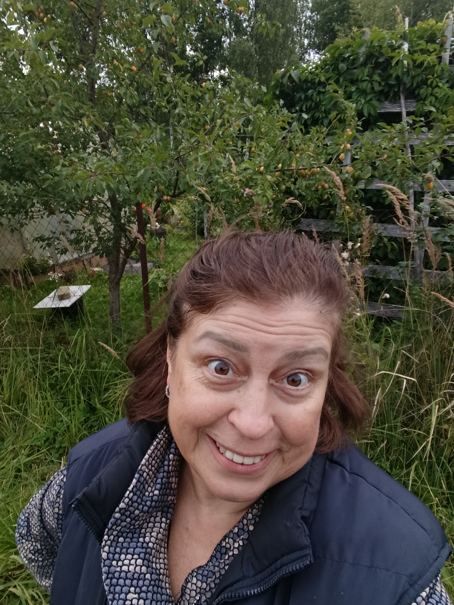 A middle-aged woman with brown hair and brown eyes is taking a selfie in a grassy outdoor area. She is wearing a dark blue vest over a patterned shirt and smiling at the camera. Behind her, there are trees with green leaves and some yellow fruit, as well as a wooden fence and tall grass. In the background, a metal object with a concrete block on top is visible, possibly part of a garden feature or equipment. The overall setting appears to be a garden or backyard in a rural or suburban environment, likely in the city of Verеya, Russia, given the context.
