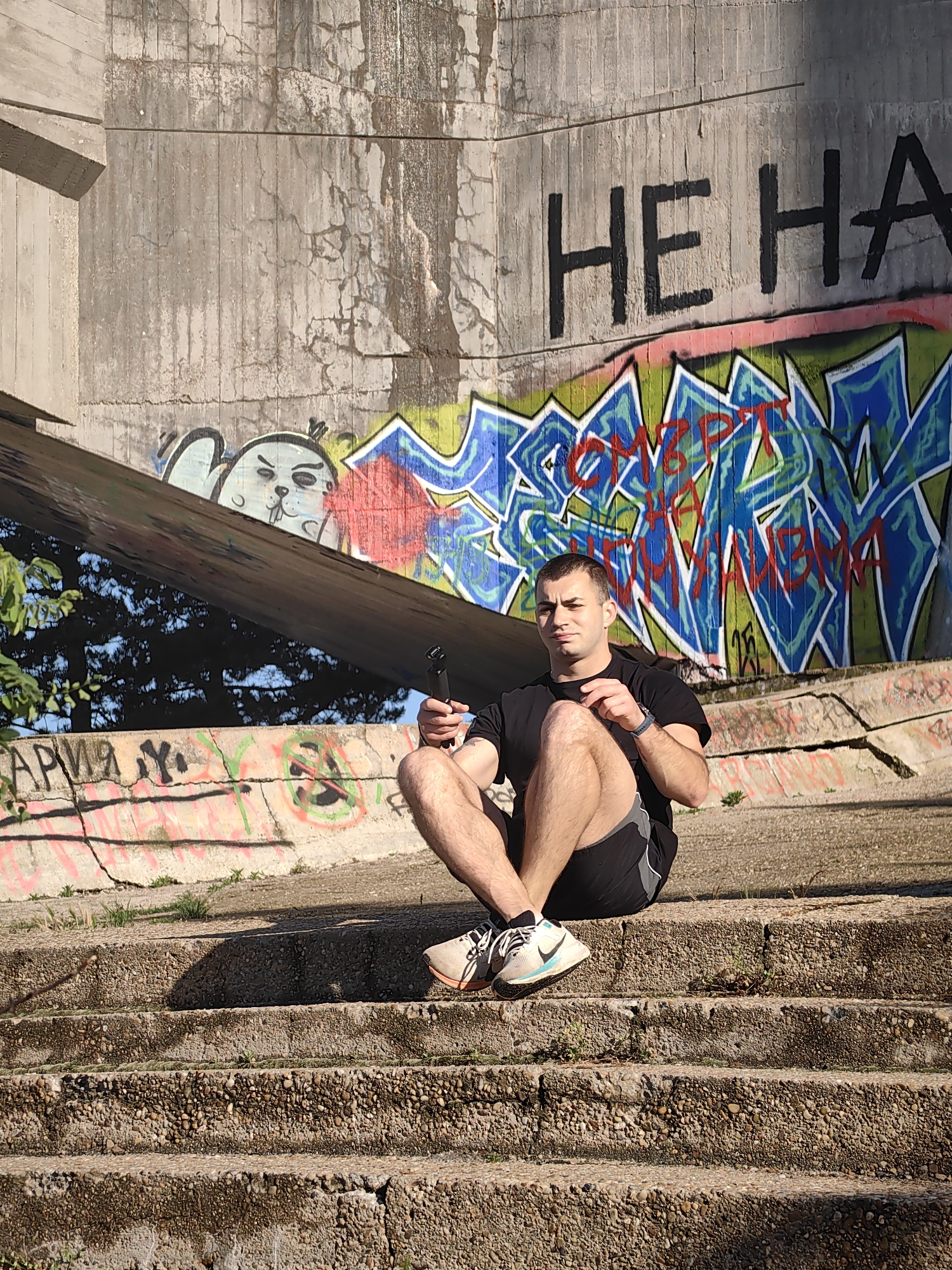 A man is sitting cross-legged on a set of concrete stairs in an outdoor location in Varna, Bulgaria. He is wearing a black t-shirt, black and gray athletic shorts, and white and blue sneakers. He is holding a black object in his hand, possibly a camera or microphone. Behind him is a concrete structure covered in graffiti, including a cartoonish cat-like face, abstract designs in blue, green, and red, and the inscription "HE HA". There are trees and sky visible under the concrete structure. More graffiti is present on the structure upon which he sits. The stairs appear to be made of weathered concrete.