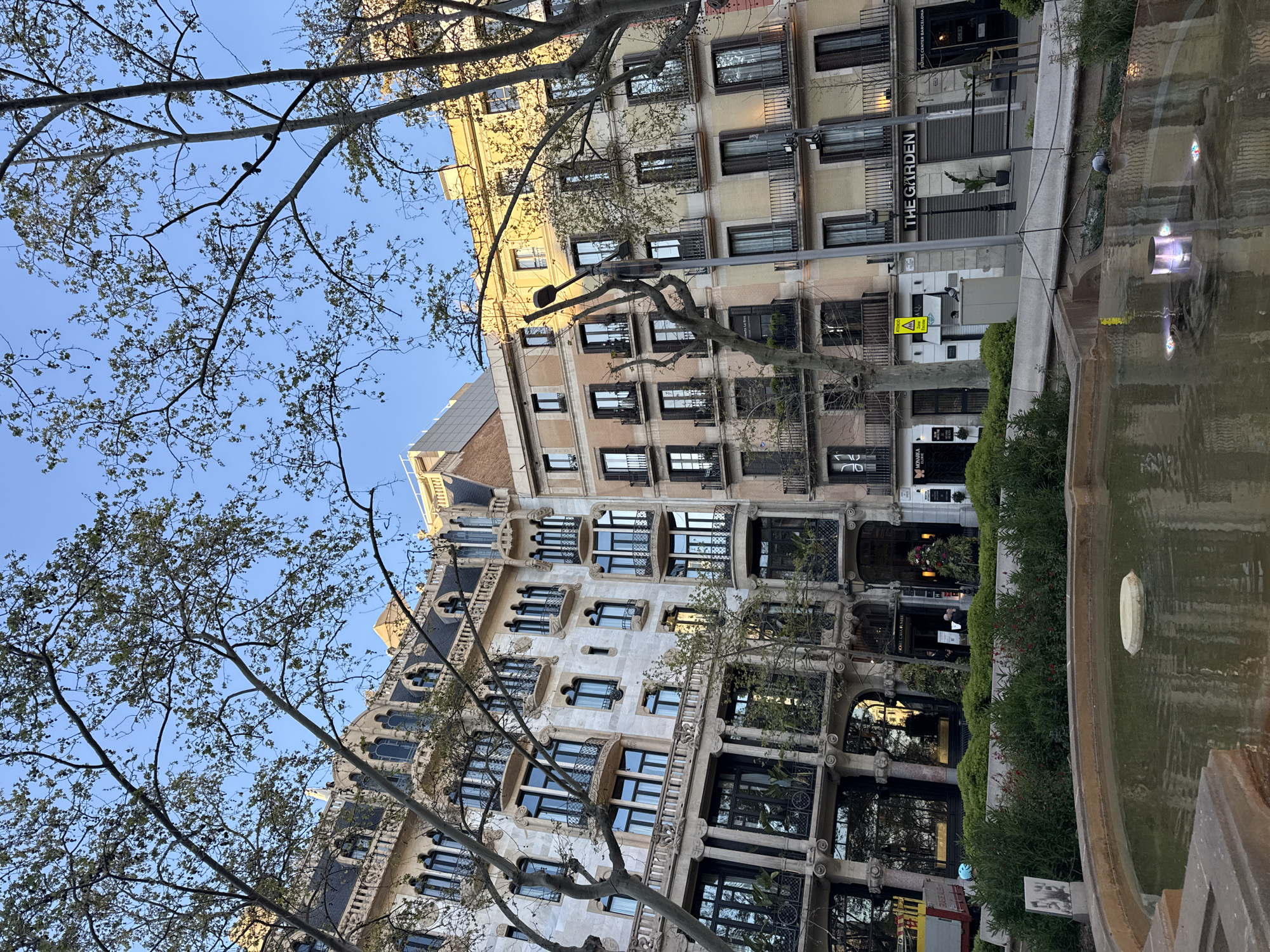 The image shows a low-angle view of a building in Barcelona, Spain, through trees and next to a water feature. The building has multiple stories with rows of windows, some of which have balconies. Its facade is adorned with architectural details, exhibiting a blend of light and dark tones, likely stone or concrete. A sign reading "THE GARDEN" is visible on the ground floor of one section of the building. A fountain with visible water is partially in the frame on the right side. Bare tree branches with budding leaves occupy the left side of the frame, reaching towards a clear blue sky. A sign with a caution symbol is visible near the base of the building. The general lighting suggests it is daytime.