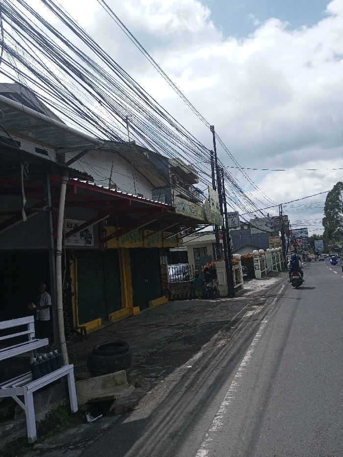 This image depicts a bustling commercial street scene. While identified by the user as Gubbisamamidi, India, the visible signage, specifically "BAKSO SOPU" and "BENGKEL" (workshop), along with the architecture, strongly indicates an Indonesian location.
The scene is under an overcast sky with white and grey clouds. A dense network of numerous black electrical and communication cables crisscrosses the upper portion of the frame, strung between multiple utility poles lining the street.
On the left side of the street, a row of commercial buildings is visible. The closest structure features a red corrugated metal awning supported by poles. Partially visible beneath this awning is a sign that reads "BENGKEL." Adjacent to it is a storefront with yellow trim and two closed dark green roll-down shutters, displaying a prominent sign for "BAKSO SOPU." In the immediate foreground on the left, a white metal shelving unit holds several dark-colored bottles, possibly lubricants. Inside the open-fronted shop to the far left, an older man in a light-colored shirt is visible, appearing to handle an object. On the ground near the storefront, two black tires are stacked horizontally next to a concrete block. Further down the sidewalk, two individuals, one wearing a head covering, are seated or crouching near a low, decorative white wall.
The asphalt road, marked by a worn white lane line, shows a person riding a dark motorcycle, wearing a helmet and dark attire, traveling away from the camera. Other vehicles, including motorcycles and cars, are visible further down the road in the distance. The right side of the road is bordered by a series of white, decorative pillars or fence sections, with tall trees visible further down the street on the right.