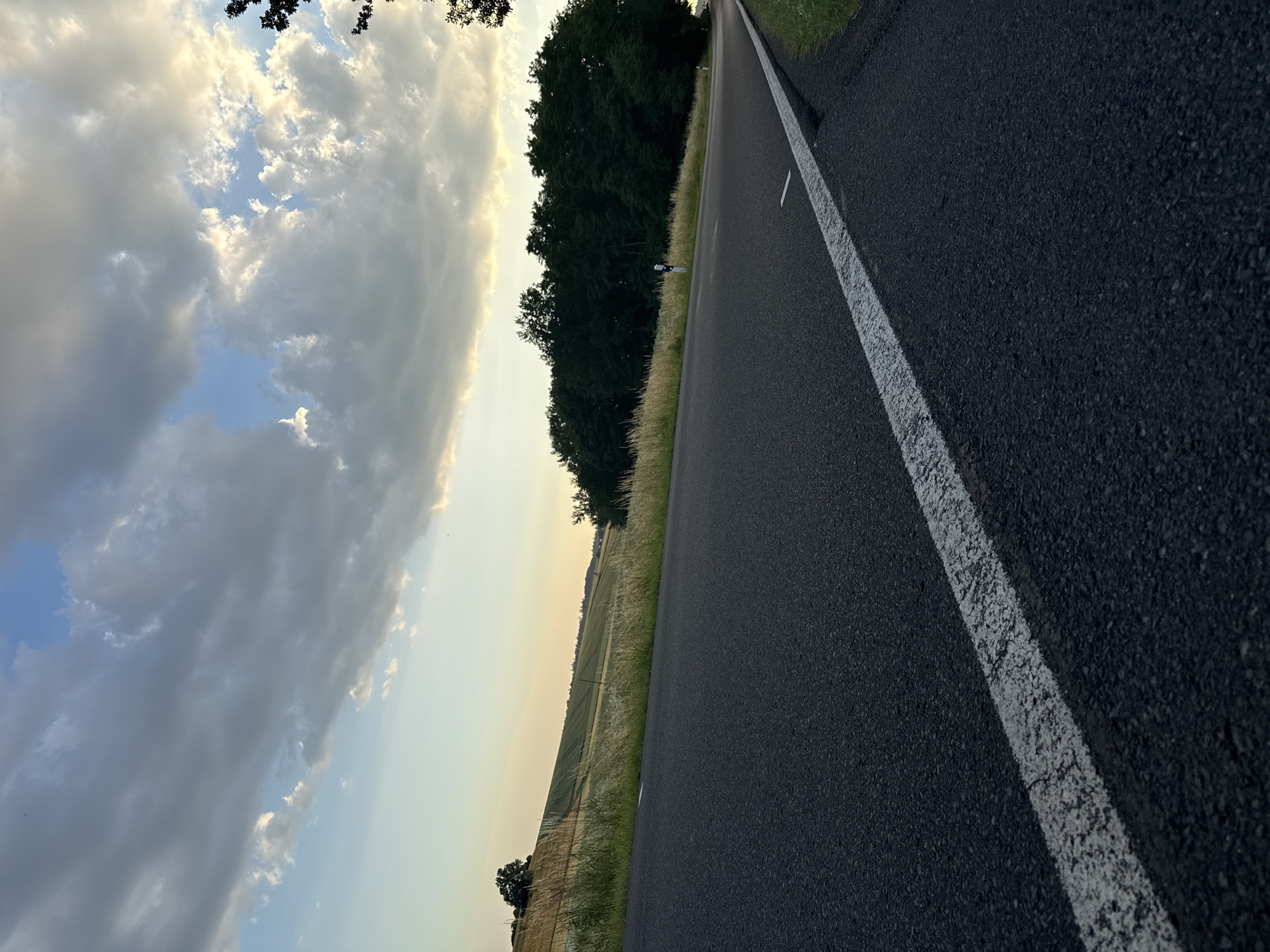 An eye-level, low-angle shot captures a paved road with a white dashed line running along its surface, curving to the right in the foreground. The road is dark gray asphalt, textured with small pebbles. Beyond the road, to the left, a gently sloping field of golden-brown crops stretches into the distance, bordered by tall, dry grass. Further back, a dense line of dark green trees forms a forest edge. Above, the sky is a mix of pale blue and dramatic, heavy gray clouds, with patches of sunlight breaking through, creating a soft, diffused light. The scene appears to be a rural setting, possibly on the outskirts of Beltershain, Germany, given the presence of agricultural land and a forest. There are no people or animals visible in the image.