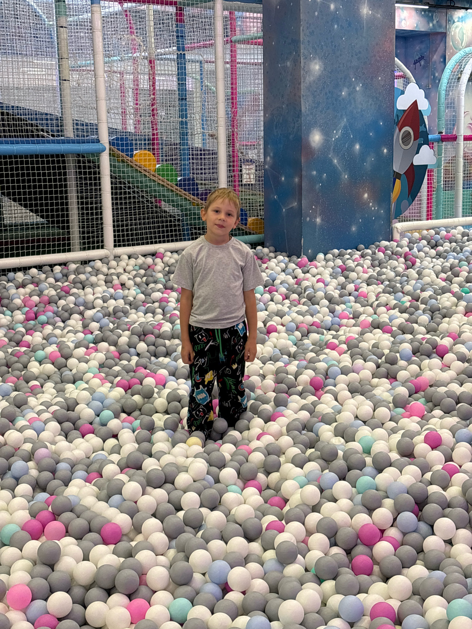 A young Caucasian boy with blonde hair and blue eyes stands in a ball pit. He is wearing a grey t-shirt and black shorts with a cartoon print. The ball pit is filled with a multitude of small, round balls in various shades of pink, grey, and white. In the background, there is a large, blue, cylindrical structure decorated with a celestial theme, featuring stars and planets. To the right of the structure, a graphic of a red rocket ship with white clouds is visible. The area appears to be an indoor playground or entertainment center, characterized by colorful netting and structures typical of such venues. The overall setting suggests a playful and recreational environment. The location is in Rusanovo, Russia.