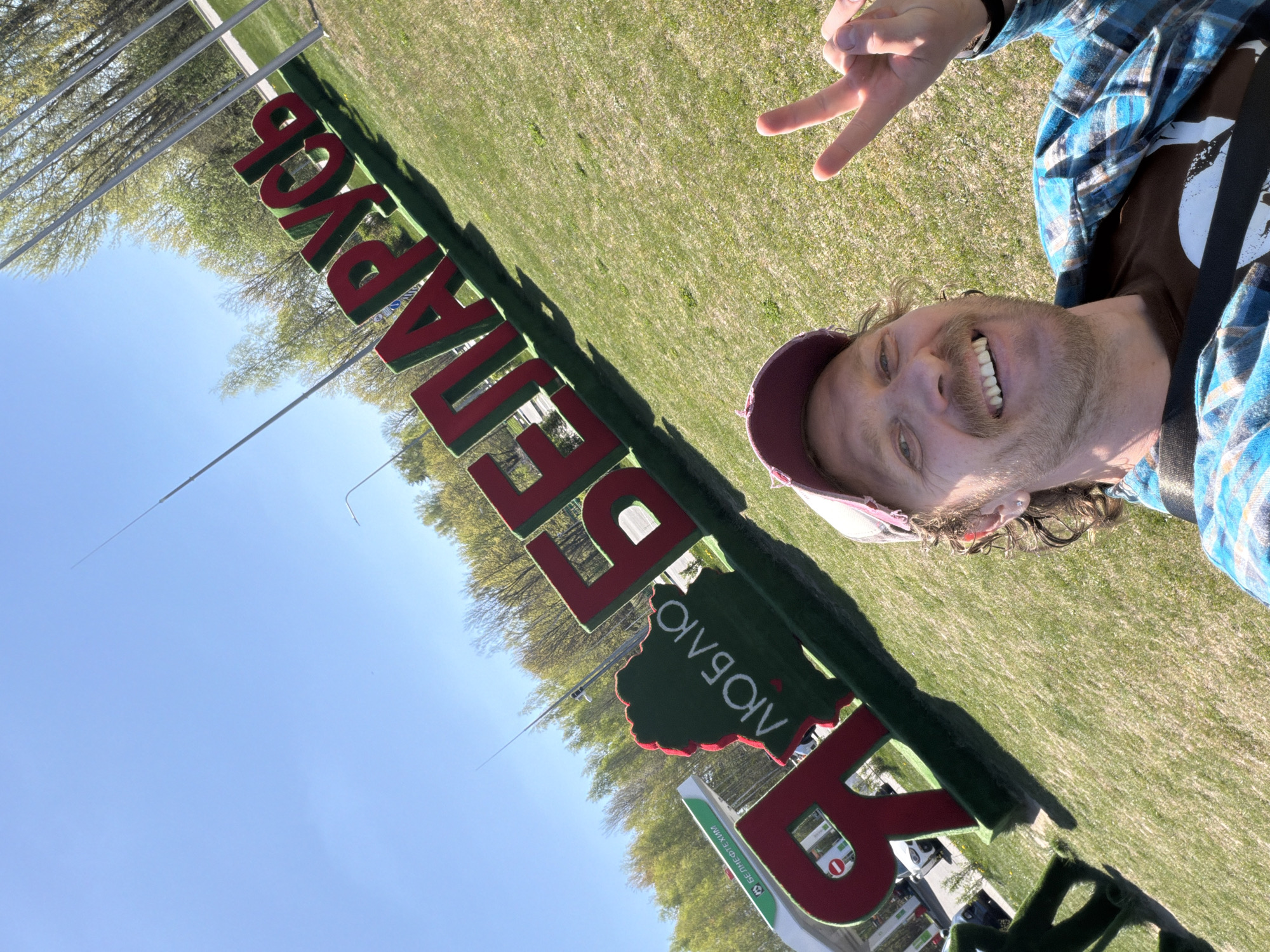 A medium shot captures a man in Крошынскі сельскі Савет, Belarus, posing in front of a sign spelling "БЕЛАГРУСЬ" (Belarus). The man is angled towards the camera, making a peace sign with his right hand. He wears a blue and white plaid shirt over a brown t-shirt, with a maroon and white baseball cap atop his head. He has light skin, a mustache, and a wide smile displaying his teeth. The sign, featuring large red letters with a green border, is prominent in the foreground. A small green sign with white Cyrillic text is visible below. The background shows a sunny day with blue sky, trees with budding green leaves, and a grassy field. Tall metal poles stretch into the sky on the left side of the frame.