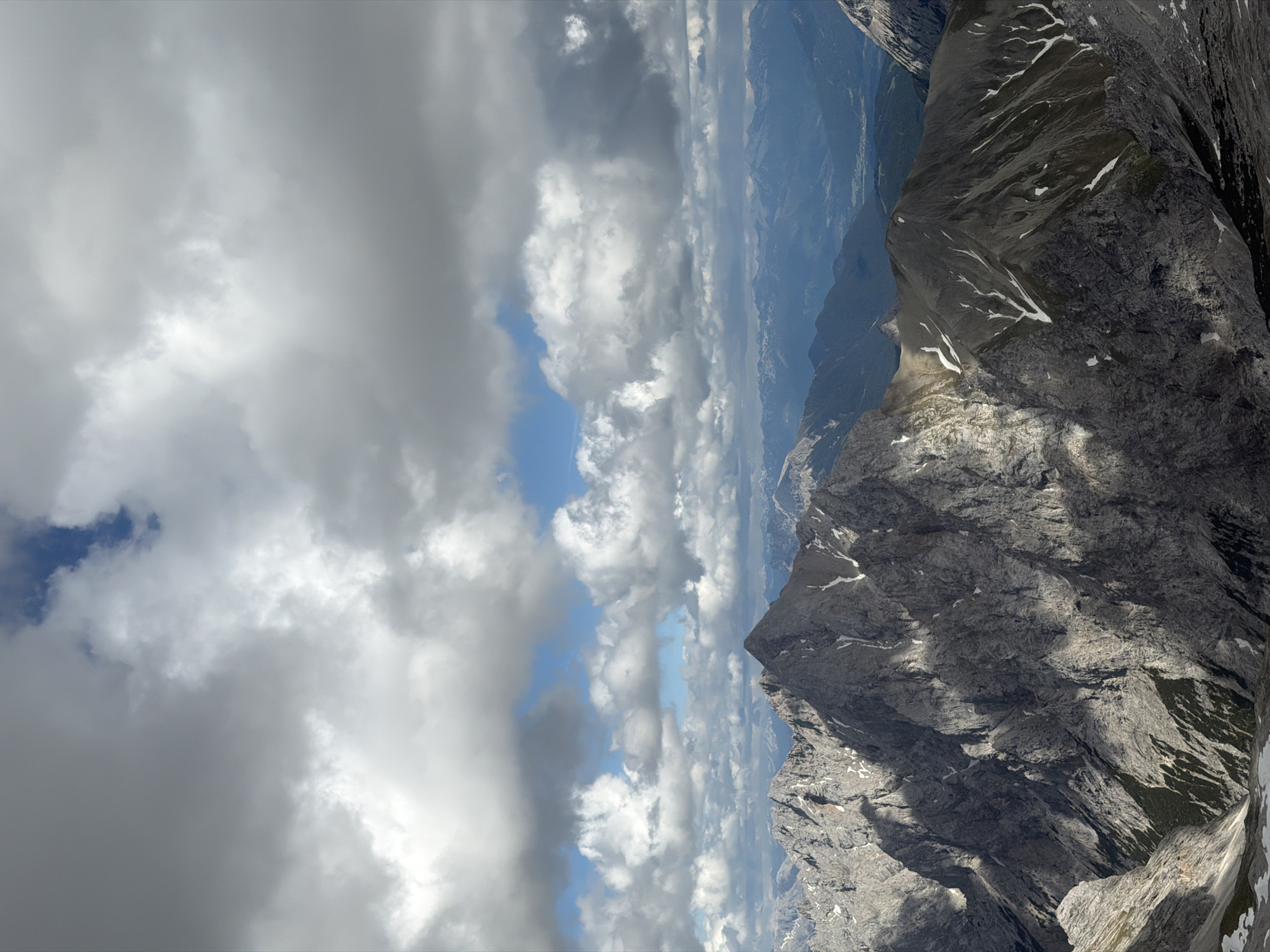 This is a high-angle, vertical shot of a majestic mountain range under a dramatic sky. The foreground is dominated by jagged, rocky peaks, with some patches of snow clinging to the slopes, suggesting a high altitude and possibly a cool climate. The texture of the rock is rough and varied, with deep shadows accentuating the ruggedness.
In the mid-ground, more mountains recede into the distance, their forms softened by atmospheric perspective. A valley or lower-lying area is visible between some of these peaks, with hints of green vegetation and possibly a settlement. The farthest mountains appear as a hazy blue ridge.
The sky above is a dynamic canvas of clouds. Large, puffy cumulus clouds are interspersed with patches of brilliant blue sky. Some clouds are bright white, indicating they are catching the sunlight, while others are darker gray, hinting at their depth and density. The interplay of light and shadow on the mountains is a prominent feature, with sunlight breaking through the clouds in places, illuminating specific features of the terrain.
While the image does not directly depict Garmisch-Partenkirchen, the scenery is consistent with the alpine landscape surrounding this Bavarian town, known for its proximity to the Wetterstein Mountains, including the Zugspitze, Germany's highest peak. This vista is representative of the breathtaking views one might experience from a high vantage point in the region. There are no people, animals, or man-made structures clearly visible in the image. The overall impression is one of natural grandeur and the powerful beauty of the mountains.