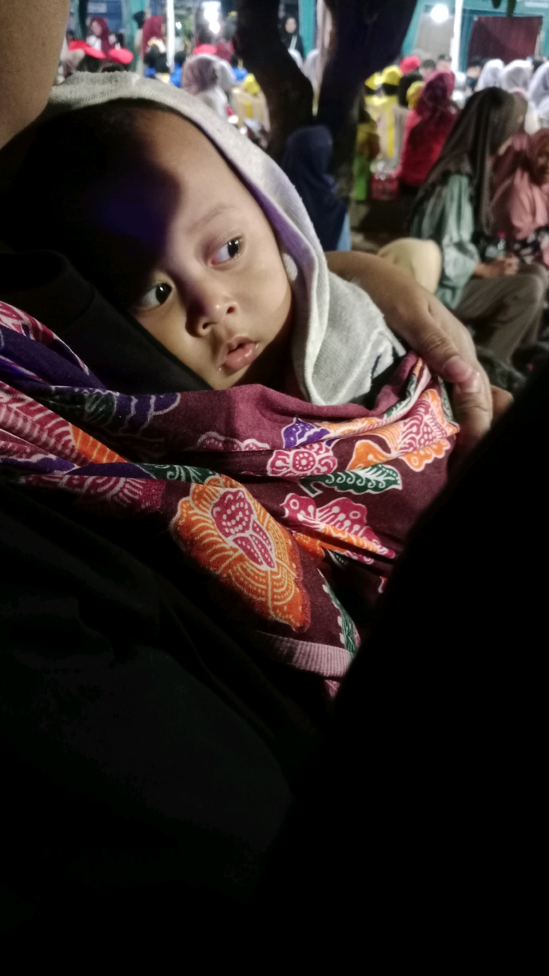 A close-up shot shows a baby with a grey hood wrapped around its head. The baby is looking to the right of the frame with a slightly open mouth. It is nestled against a person's chest, which is covered by a dark garment. The baby is also wrapped in a patterned cloth with a burgundy base and intricate designs in orange, purple, and white. The background is out of focus and appears to be an outdoor setting at night, possibly a gathering or event, with blurred figures of people in colorful clothing, trees, and structures visible. It is not possible to determine the specific location within Tambran, Indonesia.