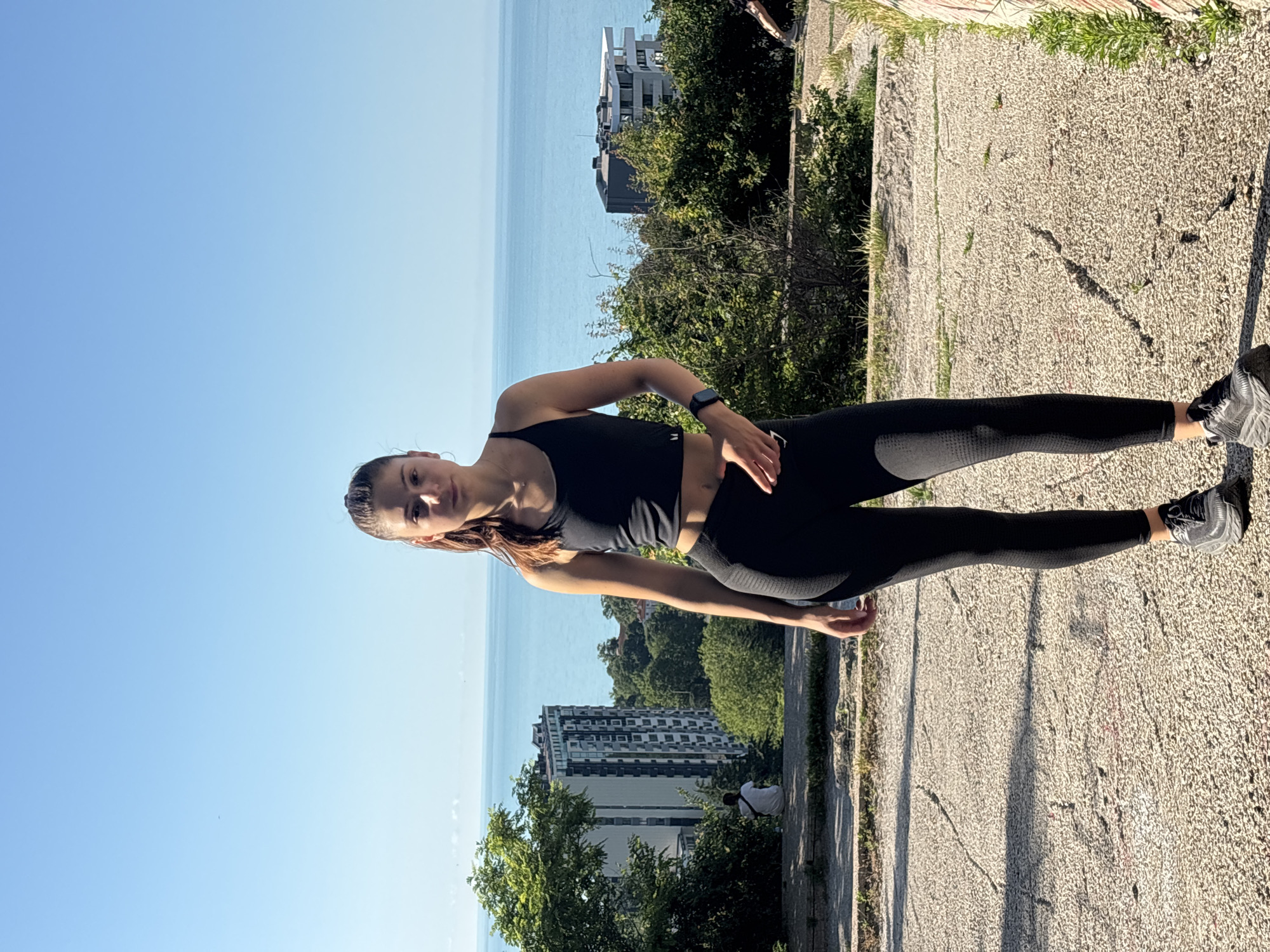 A young woman stands outdoors on a paved surface, facing the camera. She is wearing a black athletic top and black leggings, with black athletic shoes. Her hair is pulled back in a ponytail. She has a smartwatch on her left wrist. In the background, there are trees, buildings, and the sea under a clear blue sky. The scene appears to be in Varna, Bulgaria, possibly an elevated viewpoint overlooking the city and coastline.