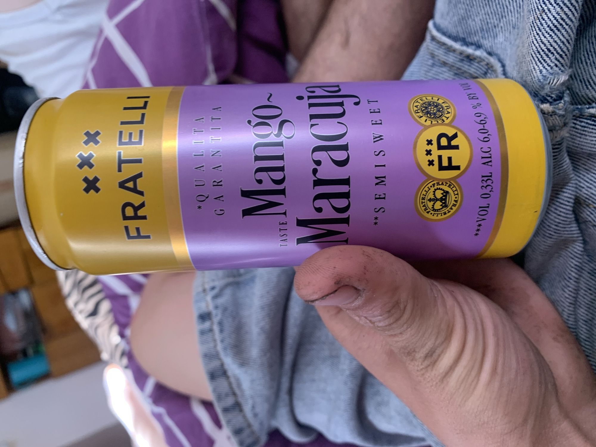 An extreme close-up shot focuses on a person's hand holding a yellow and purple aluminum can of "FRATELLI Mango ~ Maracuya" drink. The can is mostly visible, with text indicating "* QUALITA GARANTITA", "Mango ~ Maracuya", "** SEMISWEET", and "VOL 0,33L ALC 6,0-6,9 % BY VOL". Two circular logos with crowns and the word "FRATELLI" are present on the can, along with the letters "FR" in a yellow circle with three x's above it. The person's hand, with dirt under the fingernails, is prominently featured. The background shows blurred elements of what appears to be a bed with purple and white striped bedding, and a light-colored surface. The image does not contain any contextual information to place it in Zhmerynka, Ukraine.