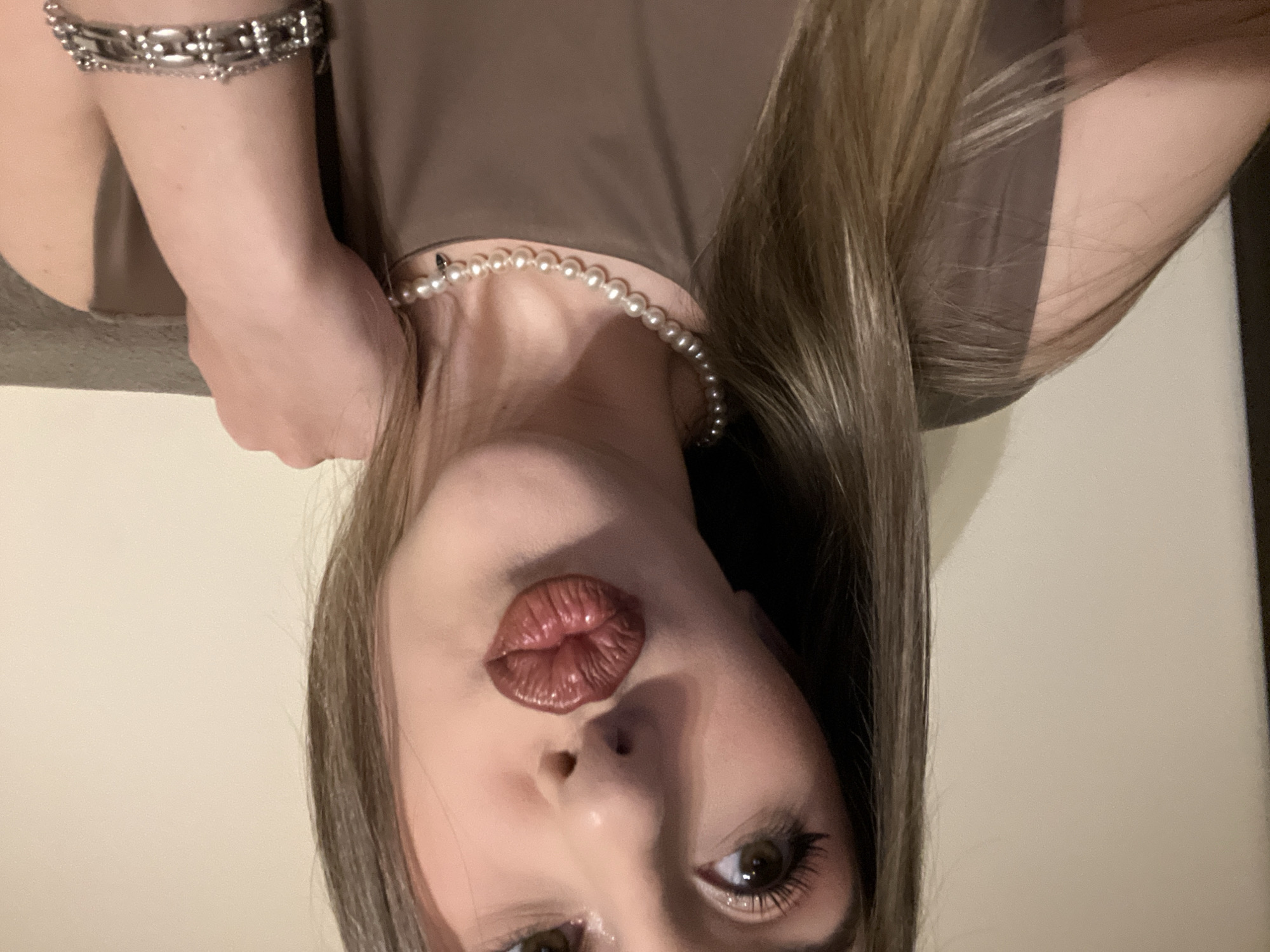 A selfie-style close-up shot depicts a woman with long, light brown hair and fair skin. She is wearing a brown top and a pearl necklace. A silver bracelet is visible on her wrist. Her lips are pursed into a kiss, with a glossy, reddish-brown lipstick. She has brown eyes and visible eyelash extensions. The background is a plain, light-colored wall. The image appears to be taken indoors. There is no specific information in the image to suggest a location in Sofia, Bulgaria.