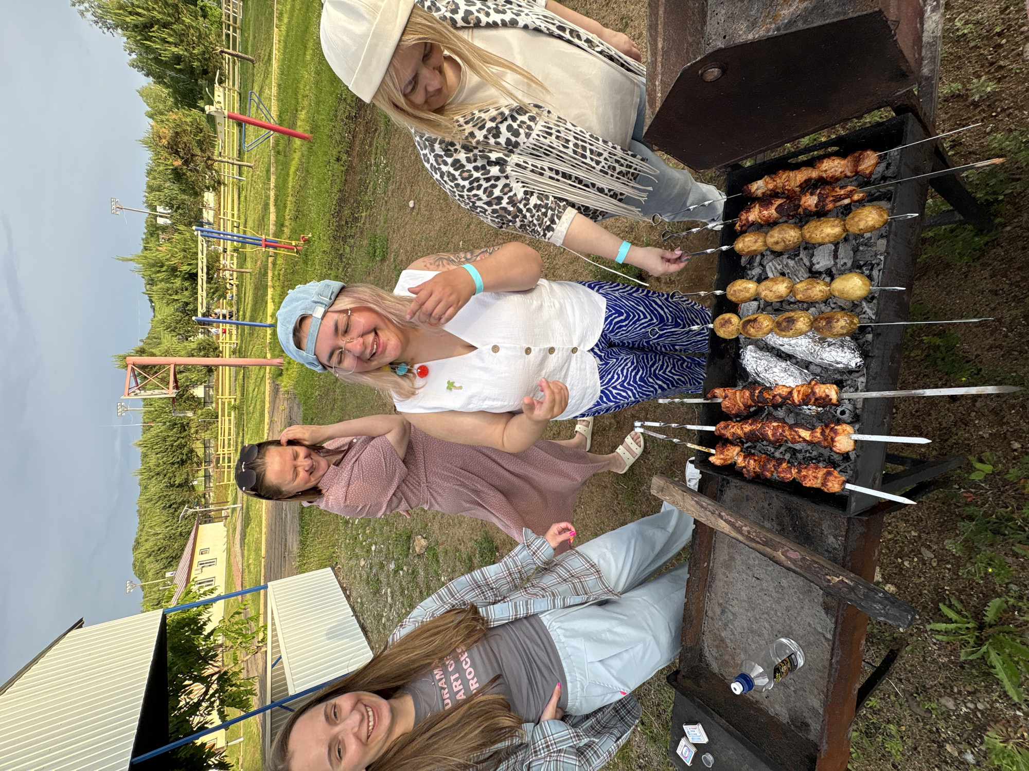 Four women are gathered around a barbecue grill in an outdoor recreational area. The woman on the far left, with long brown hair, wears a plaid shirt over a gray t-shirt and light blue pants, smiling at the camera. Next to her, a woman in a pink polka-dot dress is laughing, her hand near her face. The third woman from the left, wearing a blue cap and a white vest over a white shirt, is also laughing and gesturing. The woman on the far right, wearing a beige beanie and a leopard-print jacket with fringe, is looking down at the grill. The grill contains skewers of cooked meat and potatoes. In the background, there is a white building, a playground, trees, and a cloudy sky. The overall scene suggests a casual outdoor gathering and meal. The location is identified as a park in Birobidzhan, Russia.