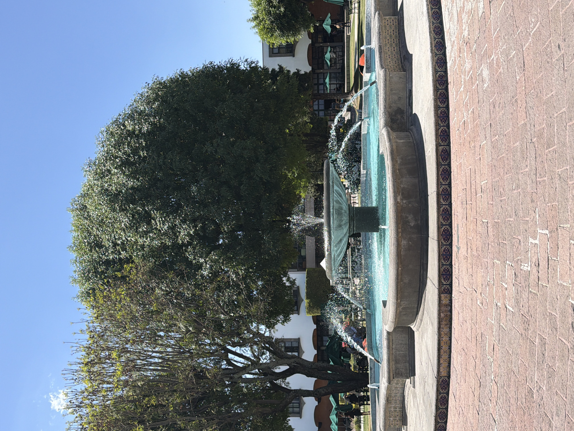 The image displays an outdoor plaza or courtyard in San Andrés Cholula, Mexico, under a clear blue sky. A multi-tiered stone fountain, centrally positioned, actively sprays blue water. The fountain's base is surrounded by reddish-brown brick paving, bordered by decorative, patterned tiles in blue, red, and yellow hues. In the background, two white, multi-story buildings with dark-framed windows are visible. These structures are partially obscured by mature trees: a large tree with sparse foliage on the left and a dense-foliage tree on the right. Below the buildings and trees, a grassy lawn contains multiple green market umbrellas sheltering outdoor tables and chairs. Several indistinct human figures are present, seated or standing beneath these umbrellas, engaged in what appears to be leisure or dining. The scene is illuminated by strong natural sunlight, casting shadows.