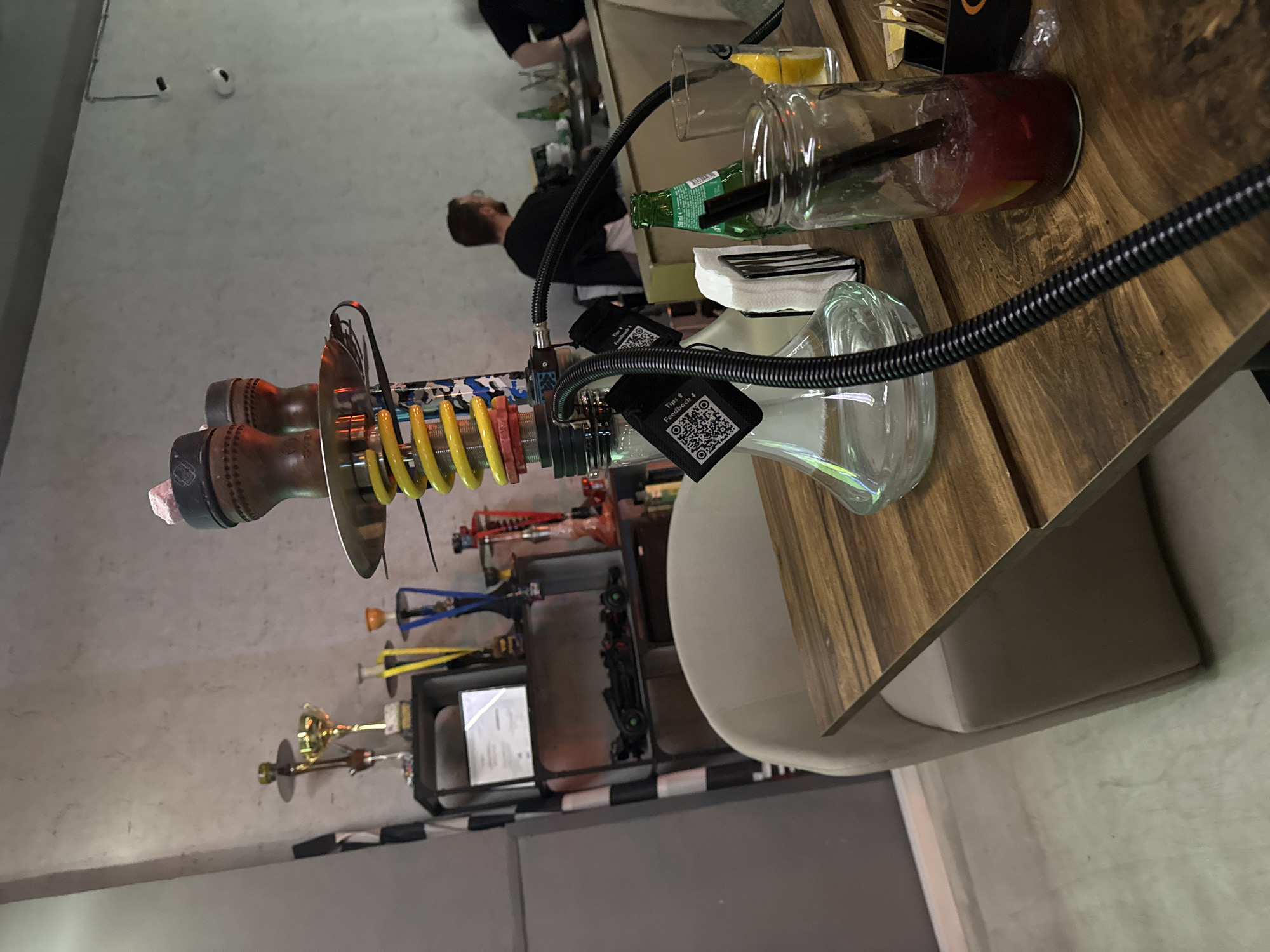 The image depicts the interior of what appears to be a restaurant or bar. There is a hookah prominently placed on a wooden table with various drinks including a cocktail, lemonade, and beer. Seated on the right, a person is partially visible, seemingly a customer. Behind the table, a man is standing at the bar; it's unclear whether he's a bartender or another patron. Additional hookahs and accessories are present on a tiered stand. The overall atmosphere suggests a modern, casual establishment. Based on the information, this is in the city of Plovdiv, Bulgaria.
