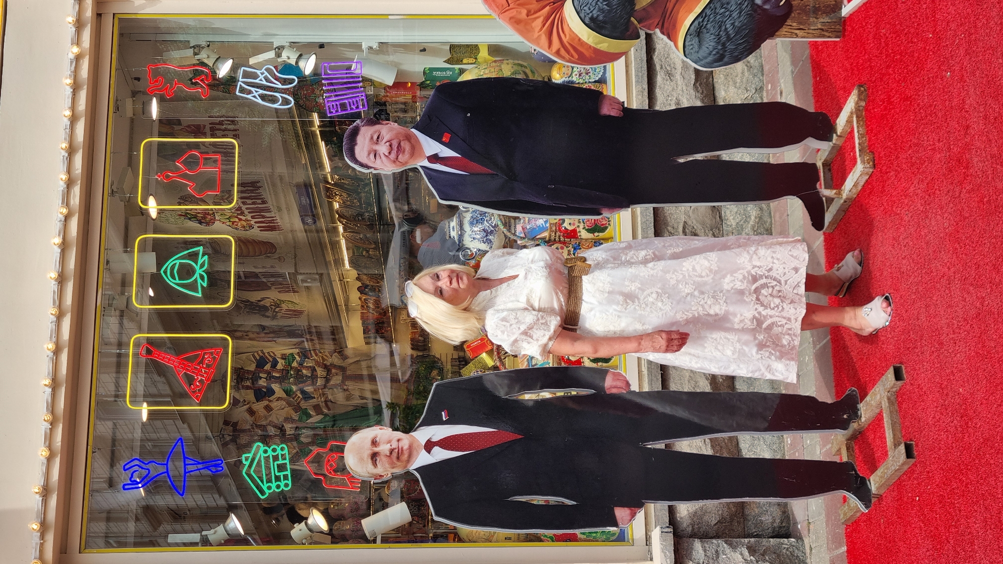 A blonde woman stands between life-sized cardboard cutouts of Vladimir Putin and Xi Jinping on a red carpet. The woman is wearing a white lace dress and light-colored sandals. Putin and Jinping cutouts are wearing suits. In the background is a shop window displaying various Russian cultural items, including matryoshka dolls, balalaikas, and traditional headscarves, illuminated by neon signs depicting similar items. The setting appears to be a tourist area in Moscow, Russia, possibly Arbat Street.