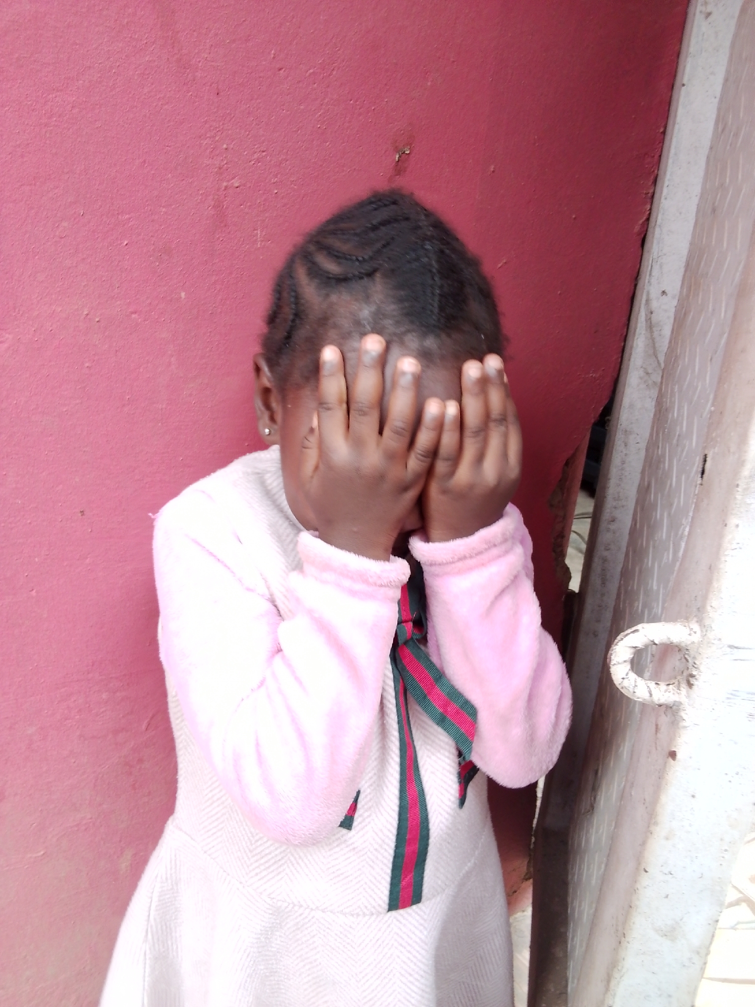 A young Black girl stands in front of a textured, dark pink wall, her face hidden by her hands. She is wearing a light pink, fuzzy sweater with a herringbone pattern on the lower half of her dress. A red, green, and black striped ribbon is tied in a bow at her neck. Her hair is styled in braids. To her right, a portion of a light-colored, weathered metal structure is visible, featuring a curved handle or latch. The scene appears to be outdoors, with natural lighting suggesting it is daytime. The setting suggests an urban or semi-urban environment, possibly in Mbar, Nigeria, given the context. The girl's gesture of covering her face implies shyness, embarrassment, or a playful game of peek-a-boo. No visible text is present in the image.