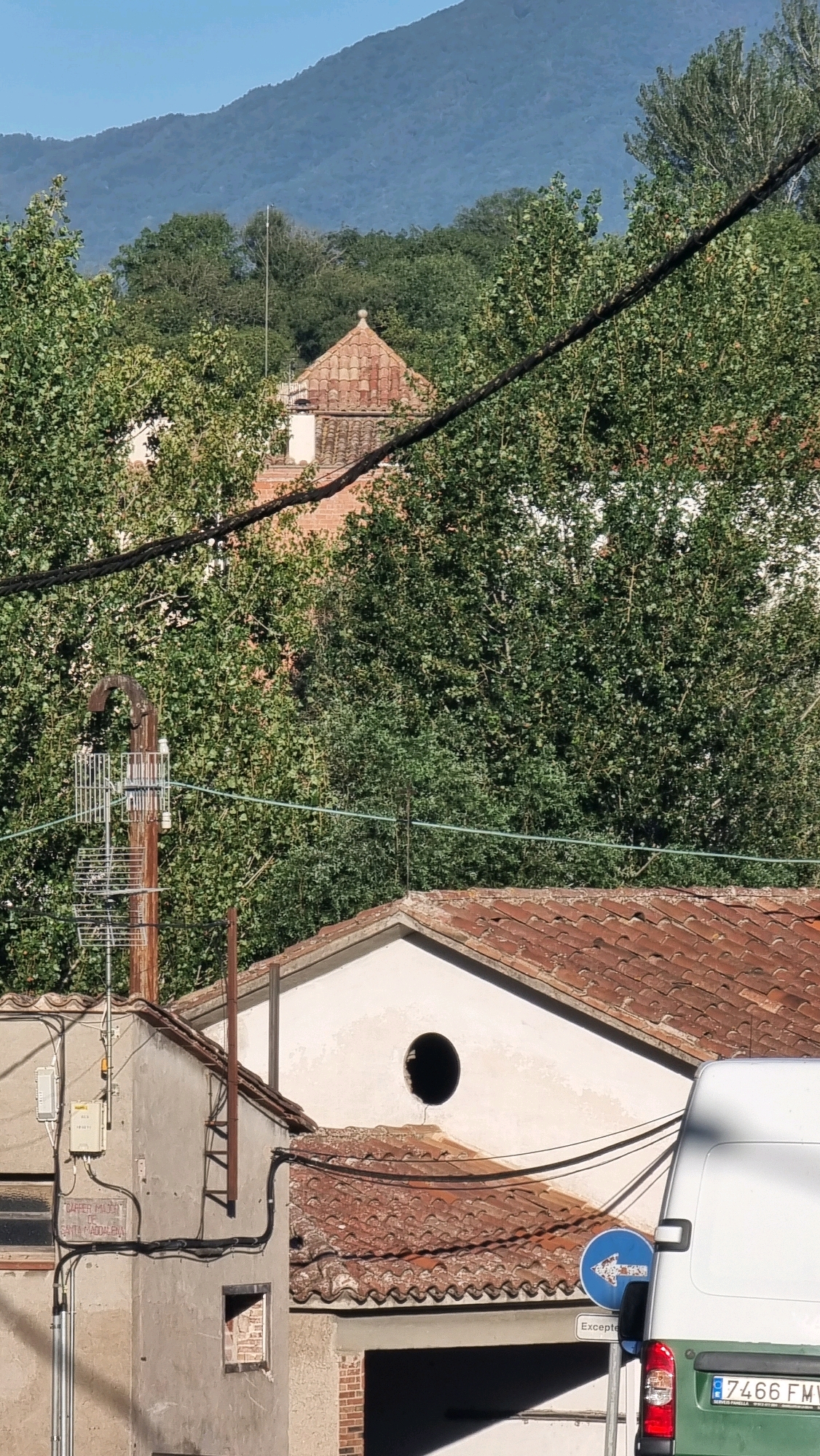 The image shows a view of buildings and trees with a mountain in the background. The scene is located in Sant Joan Les Fonts, Spain. In the foreground, there are two buildings with red tile roofs. One building has a ladder attached to its side and electrical equipment mounted on it. The other building has a circular window. A white van is parked on the right side of the image, with its rear visible. A blue traffic sign with a white arrow pointing left is also visible. The middle ground is filled with lush green trees, and a more distant mountain range forms the background. The sky is clear and blue.
