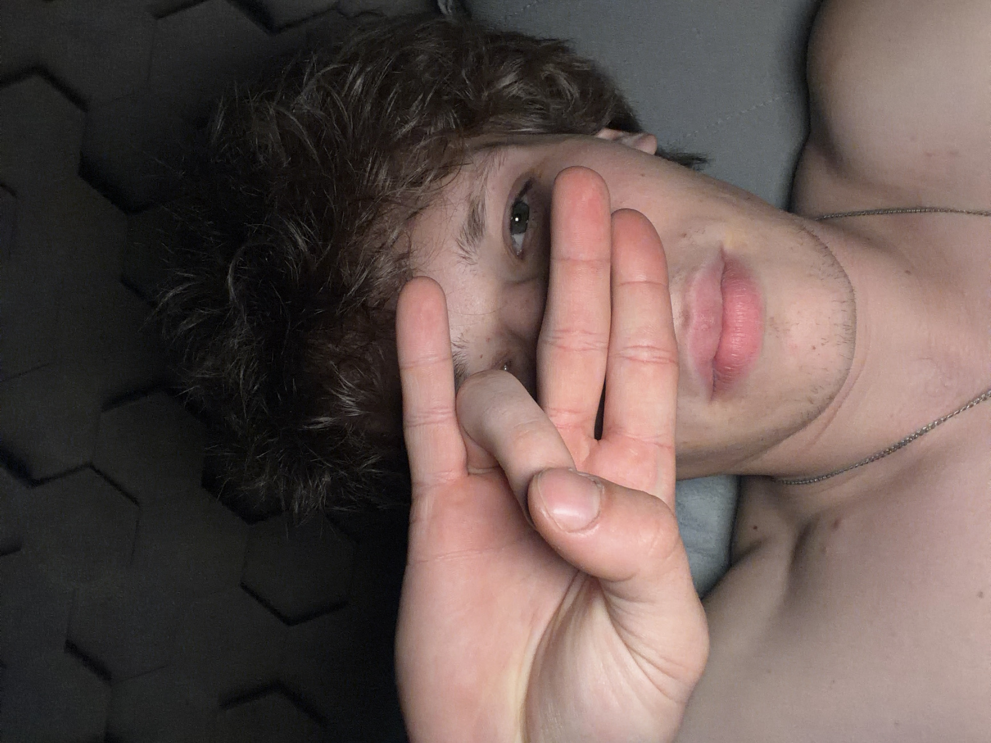 The image shows a close-up, selfie-style shot of a young, fair-skinned person with messy brown hair. The person's right hand is held up, obscuring their eyes and forehead. Their left eye, which is green, is partially visible. They are wearing a silver chain necklace. The background consists of a dark, textured surface with a hexagonal pattern. The lighting is dim and appears to be from above. The person is shirtless. There are no visible landmarks or clues to indicate the location in Nizhny Novgorod, Russia.