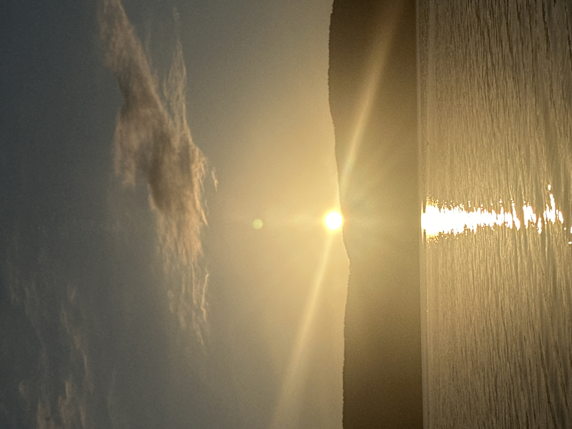 An outdoor, landscape photograph captures a sunset over the Batak Reservoir in Batak, Bulgaria. The sun, a bright orb, is positioned low on the horizon, casting a warm golden light across the scene. The sun's rays fan out, creating a visible sunburst effect. The sky above is a gradient of blues and grays, with wispy clouds tinged with orange and yellow from the setting sun. In the foreground, the calm waters of the reservoir reflect the golden light, with a prominent reflection of the sun shimmering on the surface. A dark, silhouetted line of hills or mountains forms the horizon, separating the sky from the water. There is a small, dark, rectangular object on the water, likely a boat or raft, near the center of the frame. The overall atmosphere is serene and tranquil.