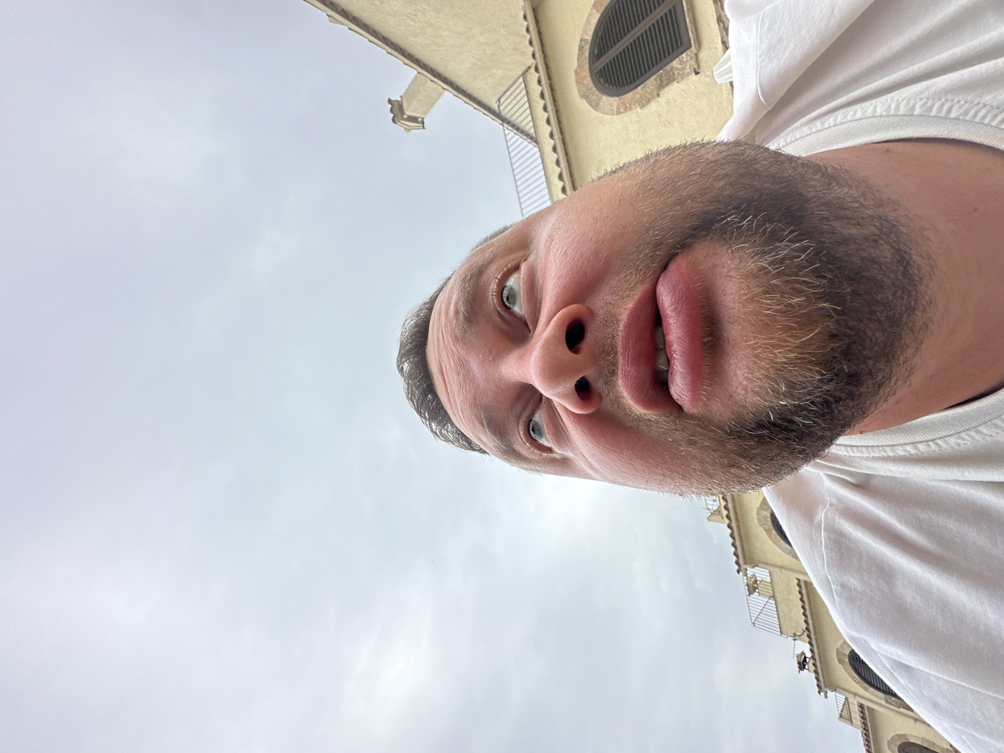 The image is a low-angle, close-up shot of a man looking upwards with a surprised or questioning expression. He has light skin, a beard, and is wearing a white t-shirt. Behind him, to the right, is a building with a pale yellow facade, a tiled roof, and arched windows with dark shutters. The sky is overcast and grey. The architectural style of the building suggests it is located in a Mediterranean coastal town. Specifically, this building is located in the town of Sant Feliu de Guíxols, Spain, as indicated by its distinctive stucco facade, terracotta roof tiles, and the characteristic arched window designs common in the region.