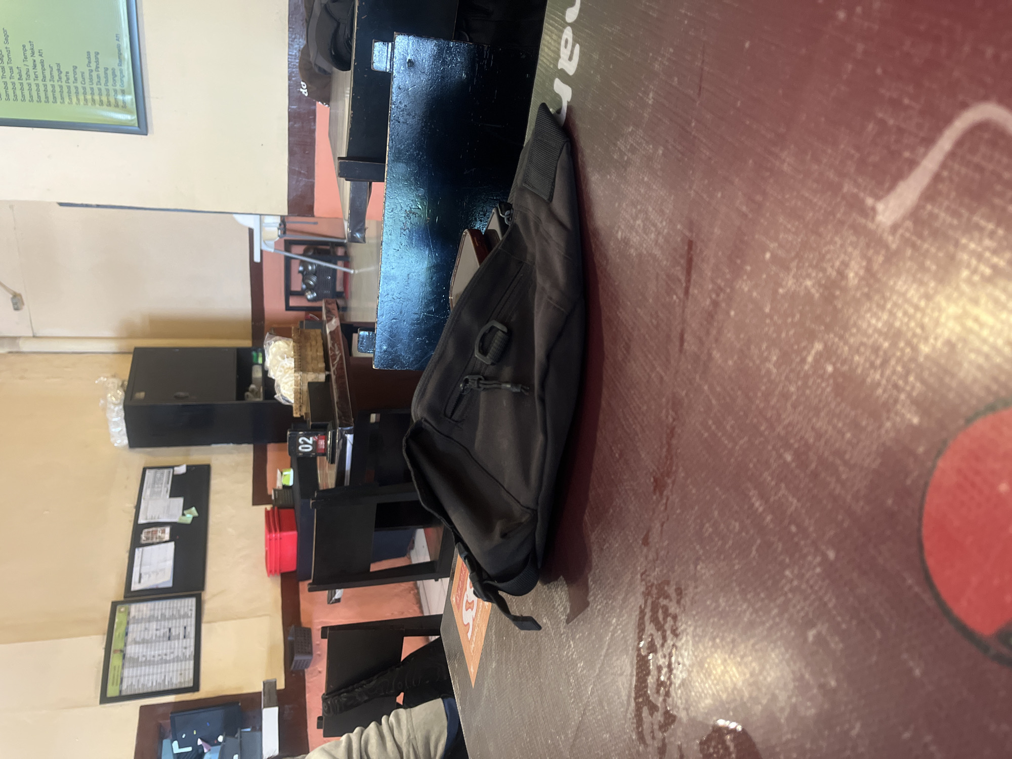 The image depicts the interior of what appears to be a casual dining establishment, possibly a warung or small restaurant, in Denpasar, Indonesia. In the foreground, a dark-colored bag, possibly a waist bag or sling bag, rests on a reddish-brown, textured tabletop. The bag is positioned at an angle, with a zipper visible and a strap extending towards the left. The tabletop shows some moisture or wetness, suggesting recent use or cleaning.
In the mid-ground, several dark-colored chairs and tables are visible. One table has a small black stand with the number "02" on it, likely indicating a table number. On another table, there are stacks of what appear to be crackers or snacks in a woven basket. A red plastic container is also visible on a table behind the chairs.
The background reveals a wall with framed items, including what looks like a menu or a schedule on the left and a corkboard with papers on the right. A tall, dark cabinet is situated to the right of the framed items. To the far right, a portion of a green menu board with text in a smaller font is visible on the wall. The overall ambiance suggests a functional, informal setting. The lighting is relatively dim, with warm tones, contributing to a cozy atmosphere. No individuals are clearly visible in the scene, although a portion of a person's arm and shoulder in a light-colored sleeve is partially visible on the left side of the image, suggesting someone was recently present at the table. The overall scene is static and unposed, capturing a moment in time within the establishment.