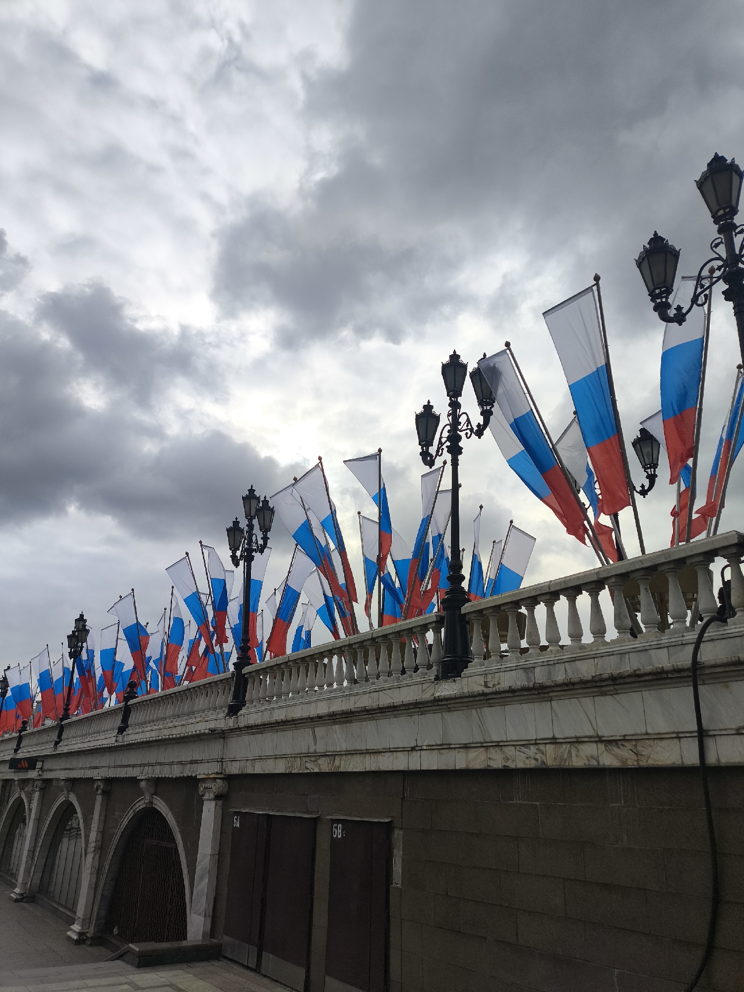 The image depicts an outdoor scene dominated by numerous Russian flags under a heavily overcast, cloudy sky. A long, ornate stone balustrade stretches across the mid-ground, adorned with a dense array of flags featuring horizontal white, blue, and red stripes. Several vintage-style black lampposts with multiple lanterns are positioned along the balustrade, interspersed among the flags.
Below the balustrade, a multi-layered architectural structure is visible, possibly part of a bridge or embankment. The upper part of this lower structure is faced with light-colored stone, featuring classical details and supporting the balustrade. Beneath this, there's a darker, block-like base with several arched openings that appear to be passages or entrances. Two dark, rectangular doors are visible on this lower section, one labeled "6A" and the other "6B".
A small, indistinct human figure can be faintly seen on the left side of the balustrade, near one of the lampposts. The scene appears to be set during daytime, but the heavy cloud cover gives a muted and somber light. The abundant display of Russian flags suggests a national event, celebration, or official decoration in a public space within Moscow, Russia.