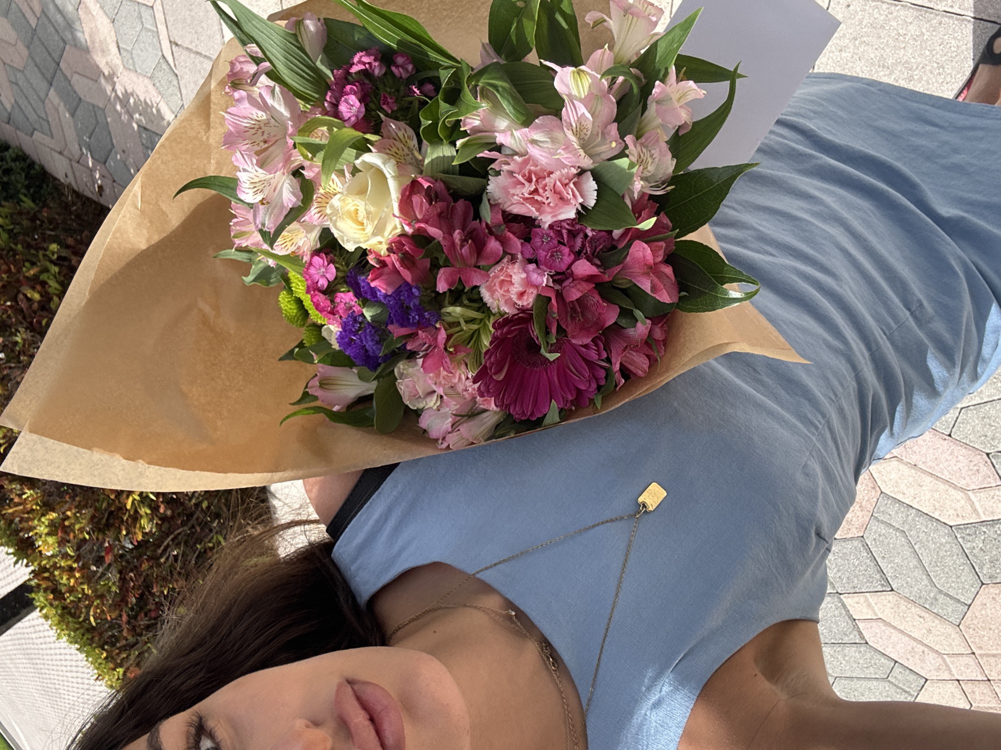 A partially visible individual is depicted from an overhead angle, holding a large bouquet of flowers wrapped in brown kraft paper. The person's lower face, neck, and upper torso are visible on the left side of the frame. She has dark hair and is wearing a light blue sleeveless dress or top. A layered gold necklace with a small charm and a rectangular gold pendant adorns her neck. The bouquet contains a dense arrangement of various flowers, including a prominent cream-colored rose, pink and white alstroemeria, various shades of pink and magenta carnations, purple statice-like flowers, a dark pink gerbera daisy, and green chrysanthemums, interspersed with green foliage. The scene is outdoors under bright sunlight. The subject is standing on a paved surface composed of geometric interlocking pavers in shades of grey and light pink. In the upper left background, out-of-focus green foliage from bushes or trees is visible. The location is Boca Raton, United States of America.