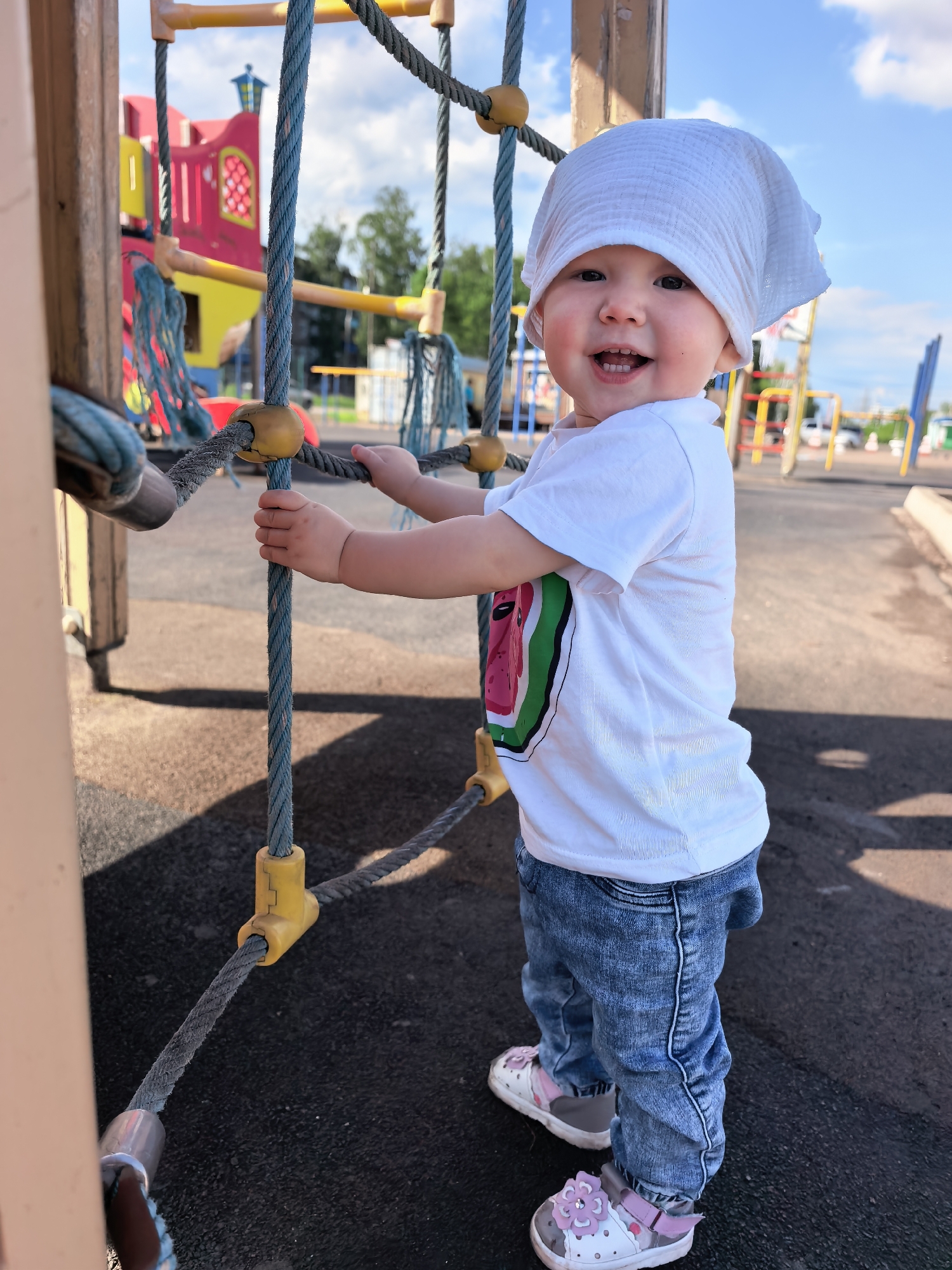 A young child is standing on a playground, holding onto a rope ladder. The child is wearing a white bandana, a white t-shirt with a watermelon design, and blue jeans. They have a happy expression and appear to be enjoying themselves. In the background, there is a colorful playground structure, trees, and a blue sky with white clouds. The ground is covered with a dark, rubbery material, typical of a modern playground. The image seems to be taken on a sunny day, possibly in the afternoon, given the lighting. The location is identified as Sterlitamak, Russia. There is no visible text in the image.