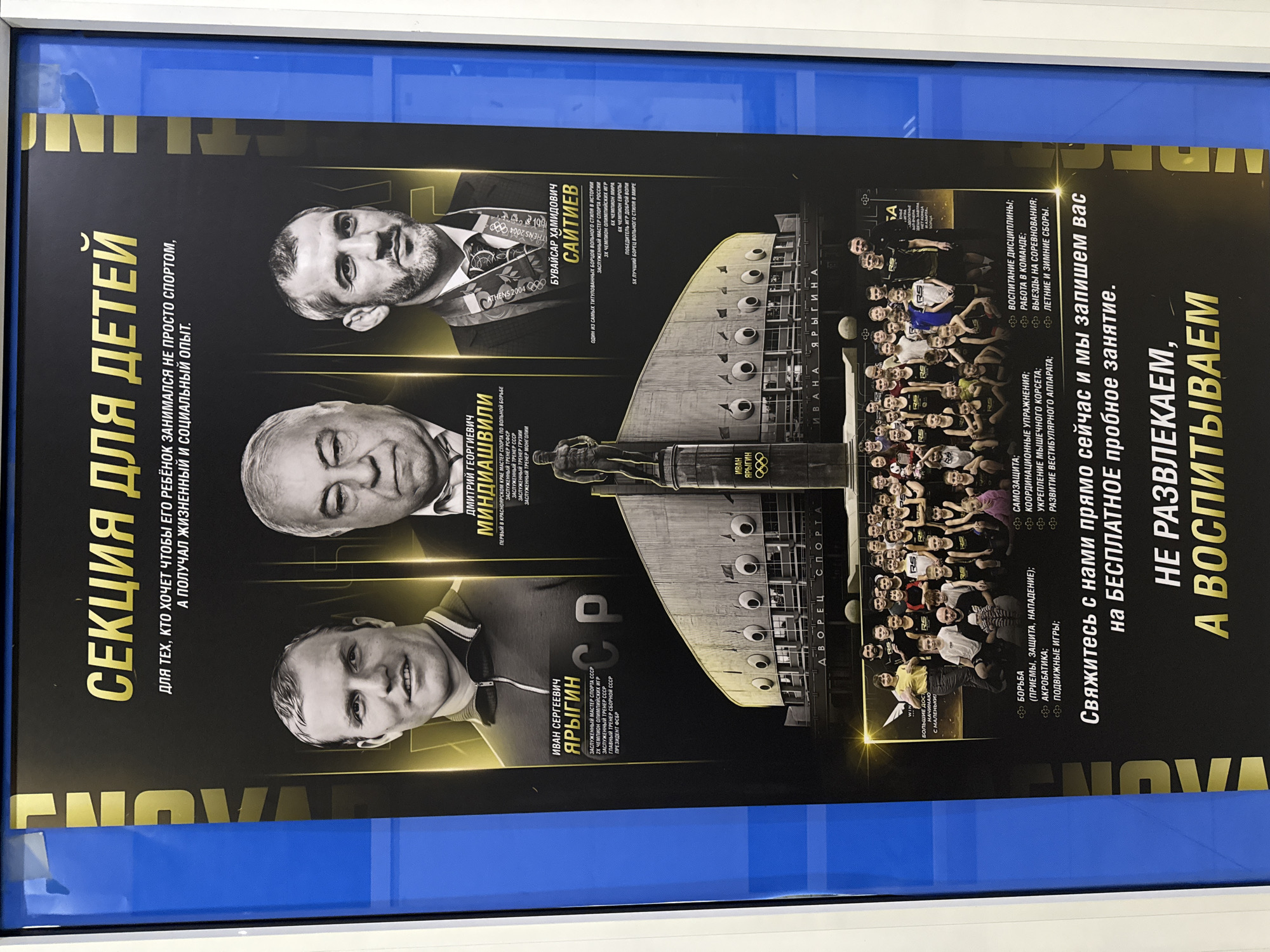 Here's a matter-of-fact description of the image:
The image shows a vertical promotional poster framed in a blue frame. The poster advertises a sports section for children, likely located in Krasnoyarsk, Russia.
The poster is divided into sections featuring portraits of three men:
1. **Ivan Sergeyevich Yarygin:** Described as an Honored Master of Sports of the USSR, a two-time Olympic champion, Honored Coach of the USSR, and President of the Federation of Wrestling.
2. **Dmitry Georgievich Mindiashvili:** Described as the first Master of Sports of freestyle wrestling in the Krasnoyarsk Territory, Honored Coach of the RSFSR, Honored Coach of the USSR, Honored Coach of Georgia, and Honored Coach of Mongolia.
3. **Buvaysar Khamidovich Saitiev:** Described as one of the most titled freestyle wrestlers in history, Honored Master of Sports of Russia, three-time Olympic champion, six-time world champion, six-time European champion, and winner of the Goodwill Games.
The poster includes a photograph of a large group of children and young adults wearing sportswear, presumably members of the sports section. Text on the poster outlines the various activities and skills taught, including wrestling techniques, acrobatics, self-defense, coordination exercises, and the development of discipline. It states that the section focuses on education rather than entertainment and invites people to contact them for a free trial session. The architecture of the Ivan Yarygin Sports Palace is also shown in the center.