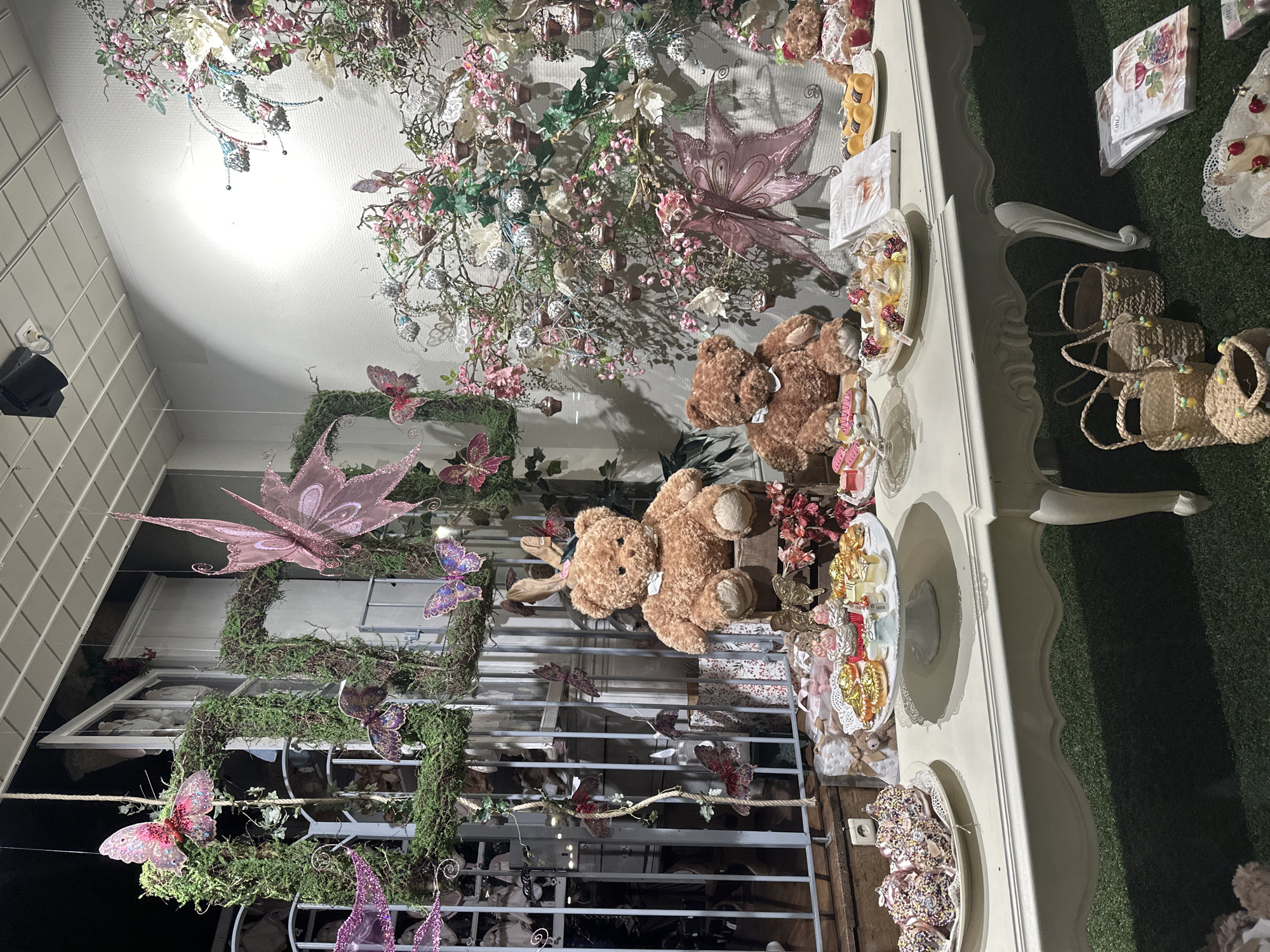 This is a detailed, forensic, matter-of-fact description of a shop window display, likely in Bruges, Belgium, featuring a whimsical, fairy-tale theme.
The scene is illuminated by soft, ambient lighting, creating a magical atmosphere. Dominating the display are two large, plush teddy bears, one wearing pink bunny ears and a white bow tie, the other with a pink bow. They are positioned as if engaged in a tea party.
In front of the bears, a long, ornate, cream-colored table displays a variety of decorative items that resemble pastries and sweets. These include small, colorful cupcakes adorned with frosting and edible glitter, gold-dusted circular treats, and what appear to be iced donuts. There are also small plates with what look like miniature cakes or tarts topped with cherries and cream. Several books or pamphlets are also visible on the table, some featuring delicate floral patterns and imagery of wine glasses and fruit.
The backdrop of the display is a lush, green, moss-covered structure forming square and rectangular frames. Draped around these frames and the surrounding area are numerous decorative butterflies in various shades of pink and purple, some with glittery wings. A large, translucent pink butterfly with intricate wing patterns is prominently featured, appearing to float in the air. Pink and white artificial flowers and branches, some with small, cupcake-like ornaments, are artfully arranged, creating a sense of an enchanted garden.
To the left of the display, a glimpse of what appears to be a building facade with a large window and railings can be seen, suggesting the shop is located within a building with traditional architecture. Some mannequins or doll-like figures are visible behind the railings.
On the ground in front of the table, several woven straw baskets are arranged, some decorated with small, colorful pom-poms and what seem to be miniature yellow lemon decorations. Further to the right, stacks of patterned napkins or paper goods are laid out.
The overall impression is that of a curated, visually rich display designed to evoke a sense of fantasy and celebration, possibly for a special occasion or a children's boutique. The presence of decorative elements such as butterflies and floral arrangements strongly suggests a connection to nature and enchantment.