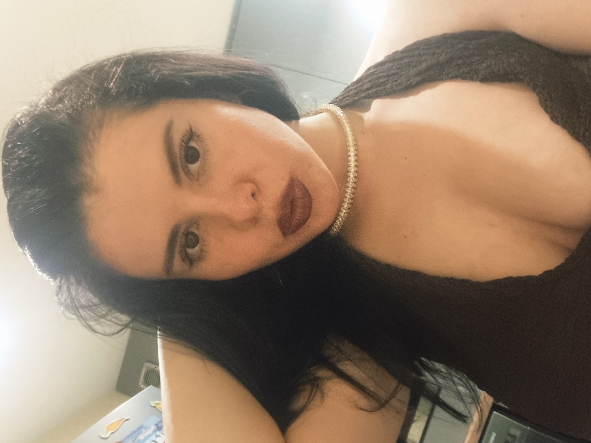 A woman with dark hair and dark lipstick is facing the camera at an angle. She is wearing a dark brown top, and a gold necklace. The background is blurred, but shows a kitchen with a refrigerator and an air conditioning unit. Location information (city of Sofia, Bulgaria) cannot be determined from the image.