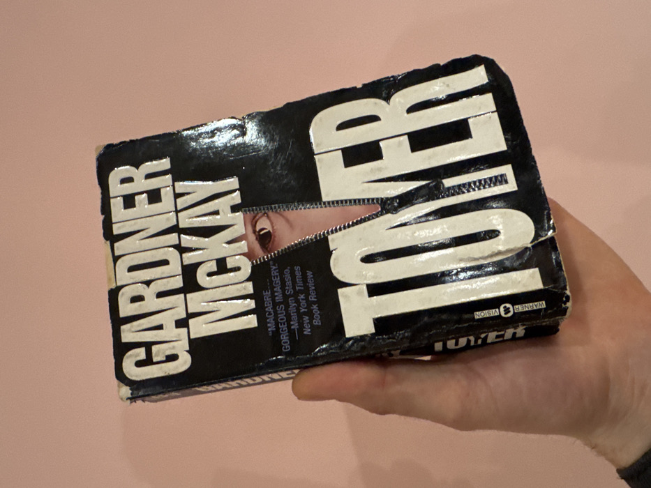 A human hand holds an aged paperback copy of the book "TOWER" by Gardner McKay, presenting its front cover against a plain, light pink background. The book's cover is primarily black with the author's name, "GARDNER McKAY," and the title, "TOWER," printed in large, white, sans-serif capital letters. A prominent design element on the cover is a vertical, black zipper motif, which appears to be unzipped, revealing a segment of a human eye (specifically the brown iris and pupil of a right eye) through the opening. To the left of the revealed eye, a quote is printed in white: "'MACABRE... GORGEOUS IMAGERY.' -Marilyn Stasio, New York Times Book Review." The book exhibits significant wear, including creasing, scuffing, and minor damage along the edges and spine. A vertical "WARNER VISION" logo is visible on the left edge of the cover. The hand, a light-skinned human hand, grips the book from the left side, with the thumb and fingers visible. A dark fabric cuff or sleeve is partially visible at the bottom left of the image. The background is a smooth, uniformly colored light pink surface, suggesting an indoor environment. This scene is located in the city of Tel-Aviv, Israel.