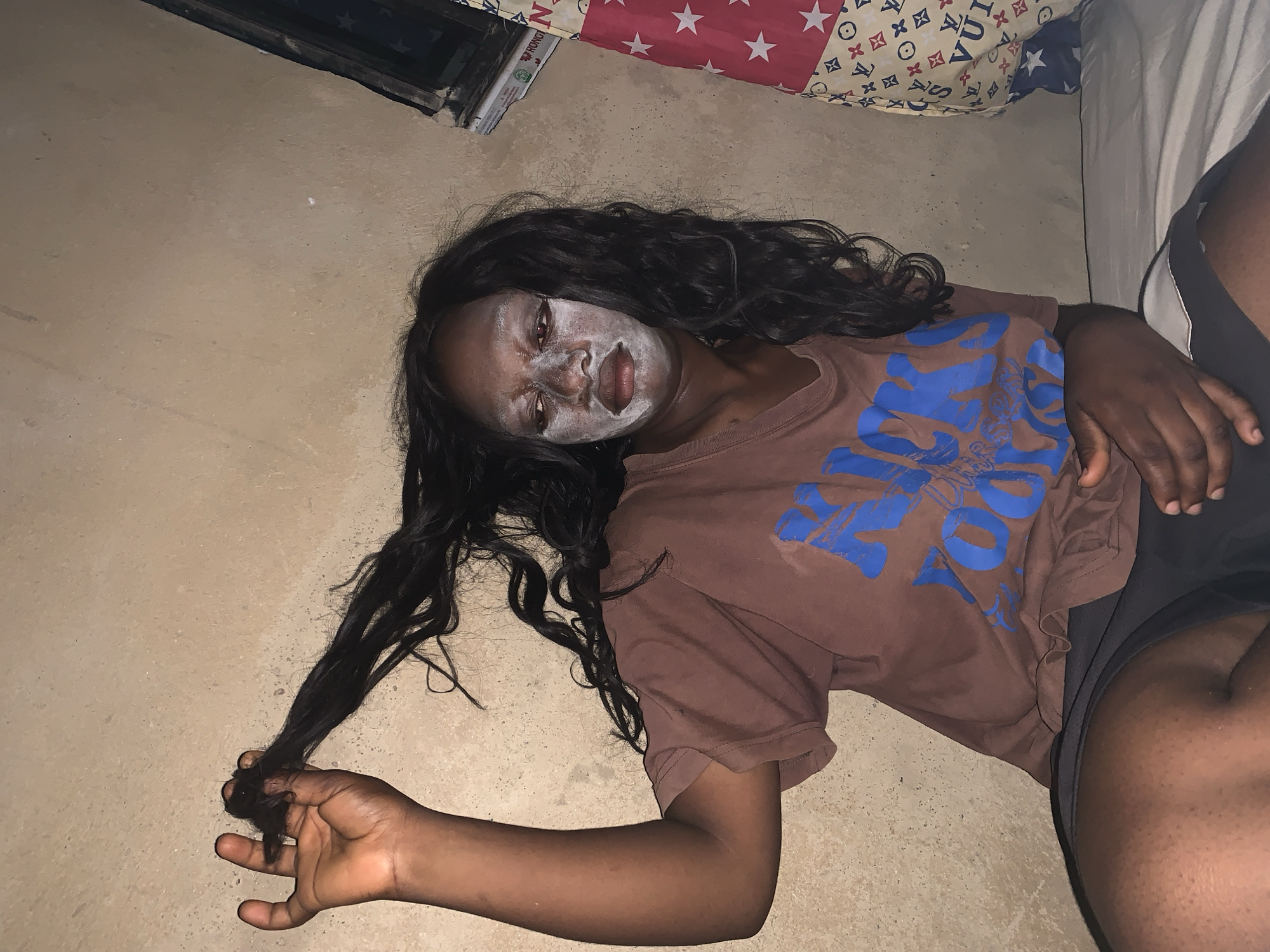 A young woman lies on a bare floor, possibly indoors, in Agban, Nigeria. She is wearing a brown t-shirt with blue lettering and black shorts. She has a white substance, potentially a face mask, applied to her face. Her long, dark hair is spread out around her head, and she is holding a section of it in her left hand. Part of a bed with patterned bedding is visible in the background.