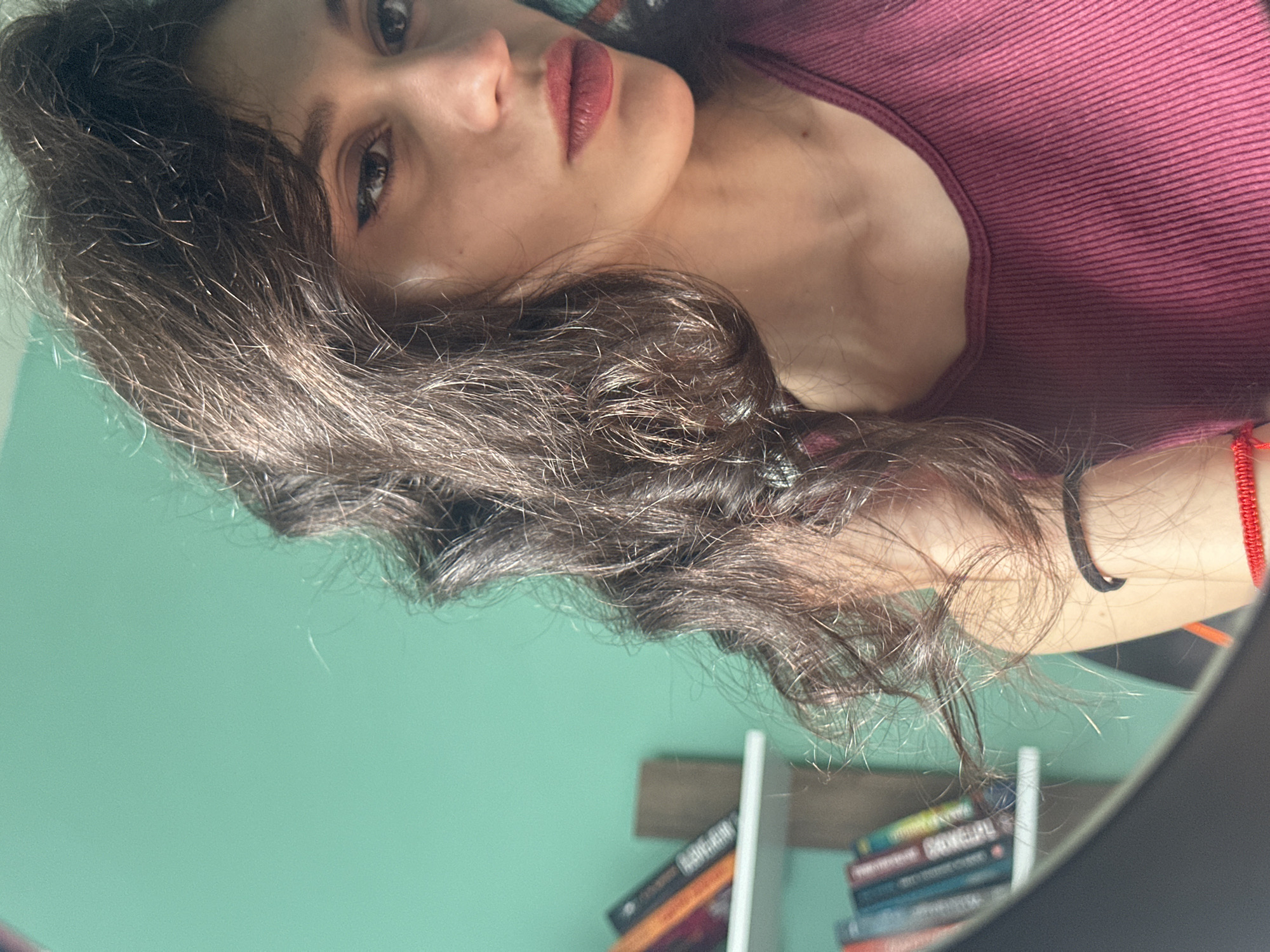 This media file is a close-up, vertically oriented selfie of a young woman. She has dark, wavy hair that partially obscures her face. Her left eye is visible, looking towards the camera, with a hint of winged eyeliner. Her lips are painted a muted berry shade. She is wearing a dark pink, ribbed tank top. A thin red string bracelet is visible on her right wrist.
The background is a bright, mint green wall, suggesting an indoor setting. A mirror is present in the foreground, reflecting the scene and adding depth. Below the mirror's reflection of the wall, shelves with stacks of books are visible. One book spine shows some text, appearing to be in a Latin alphabet, possibly in English or a similar language. The lighting suggests it is daytime, with sunlight casting highlights on her hair and skin. The overall mood appears casual and intimate. The metadata indicates the location is Sofia, Bulgaria.