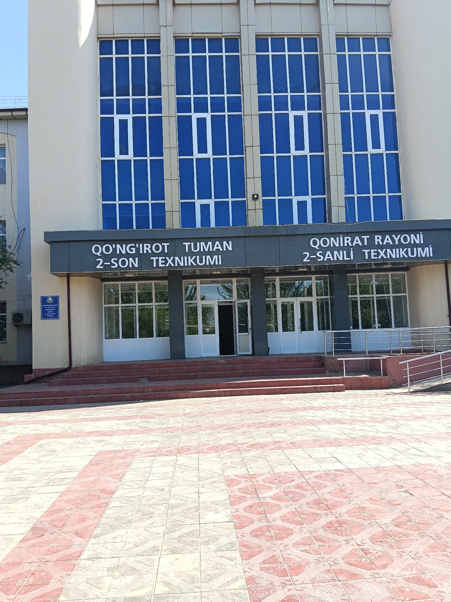 This is a frontal exterior shot of a modern building in Kungrad, Uzbekistan, featuring large blue-tinted windows with white framing on the upper floors and glass doors with white framing on the ground level. Above the entrance, a dark gray sign displays text in white lettering. The building has a light beige facade and is accessed via a wide set of red brick stairs leading to the entrance. A paved walkway with a patterned design of red and beige tiles stretches out in front of the stairs. To the left of the entrance, a blue plaque with crests and text is affixed to the wall. A metal railing system guides people up a ramp to the right of the stairs. The sky is visible in the upper left corner of the image, and some greenery can be seen through the glass doors. The overall impression is one of an institutional or educational facility.