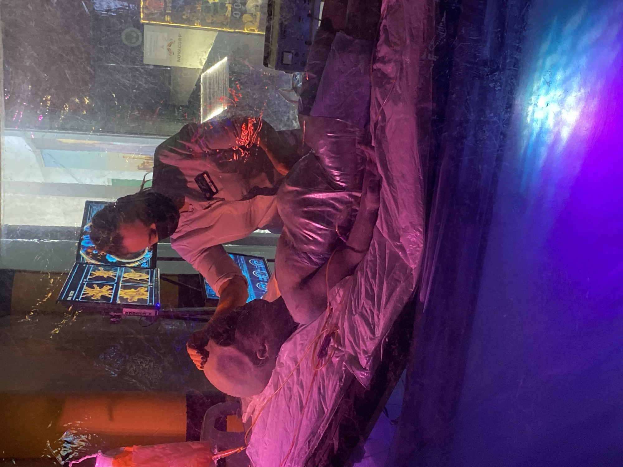 The image depicts a medical simulation or display, likely within an exhibition or museum. A mannequin lies on a white, draped surface, seemingly a medical bed. A person, likely a demonstrator or another mannequin, is positioned over the mannequin, appearing to perform a medical procedure. Medical monitors displaying graphic visualizations are visible. The environment is artificially lit with purple and blue hues. "Novacorp" is written on a small sign above them, suggesting this is either a fictional universe or themed experience. Based on the details, and in particular the mention of "Novacorp" in conjunction with what looks like a recreation of a medical simulation, this is likely a display at a science museum or science center somewhere in Yekaterinburg, Russia.
