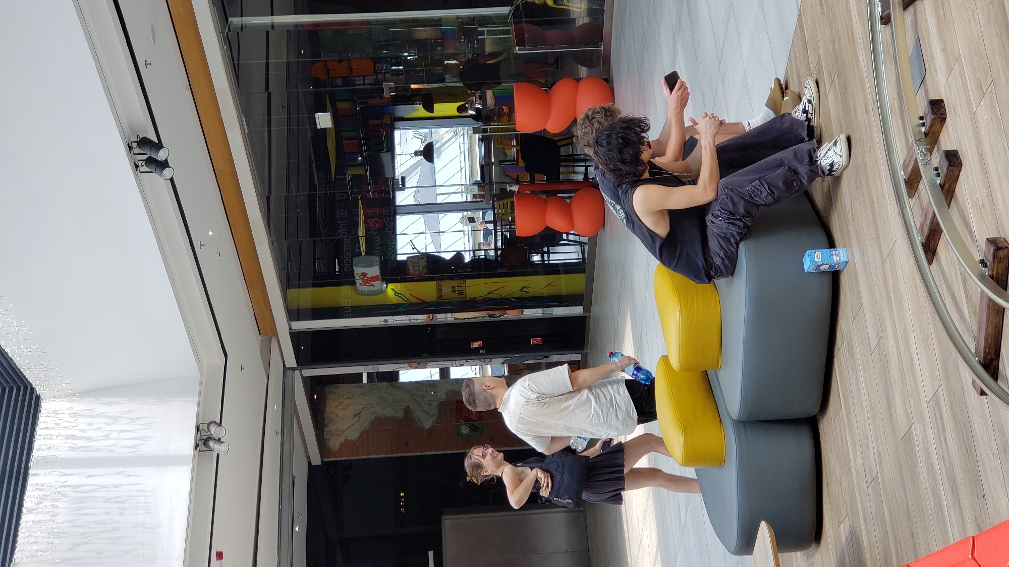 The image captures a scene in a modern indoor public space, possibly a shopping mall, given the presence of shops and seating areas. The orientation of the photo is rotated.
In the foreground, a young couple is seated on a modular sofa. The man, with dark, curly hair and wearing a black tank top and distressed jeans, is looking at his phone. Beside him, a woman is also looking at her phone. They are positioned on a grey sofa with bright yellow accents. A small blue box with cartoonish imagery is placed on the sofa.
Further back, another couple is also seated. The man wears a white t-shirt and dark shorts, holding a plastic water bottle. The woman, with blonde hair tied back, is wearing a dark top and skirt, and appears to be smiling.
Behind the seating area, a shop with colorful displays and reflective glass windows is visible. The shop's interior is filled with shelves displaying various items, and there are bright orange, mushroom-shaped stools. The architecture suggests a contemporary design with clean lines and ample lighting. The floor is made of light-colored wood planks. The time of day appears to be daytime, with bright, diffused light indicating an indoor setting.
There is some visible text in the background, including what appears to be a "Wi-Fi" sign on a yellow banner in one of the shops. The overall atmosphere seems relaxed and casual, with people taking a break and using their mobile devices.