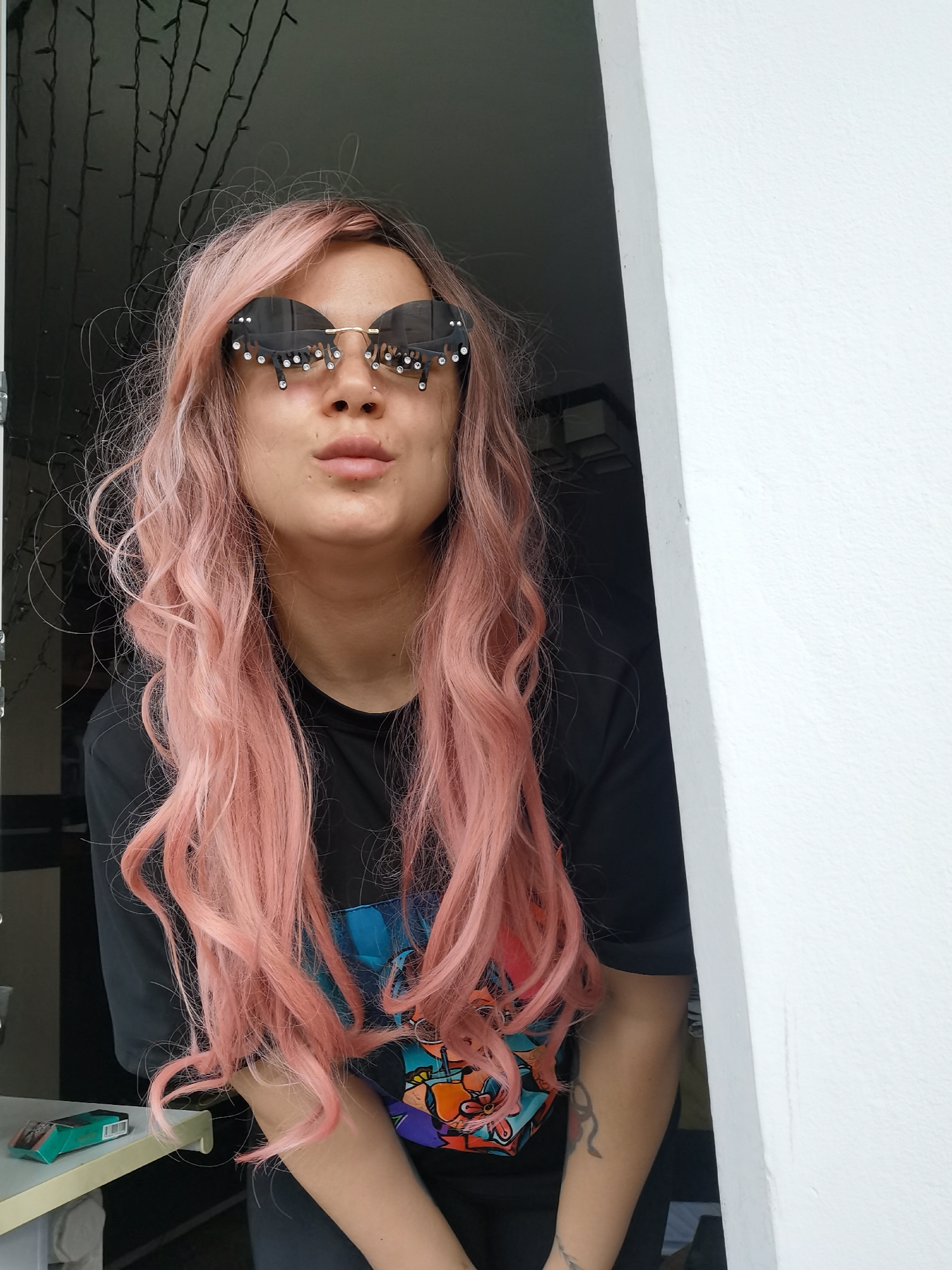 A young woman with long, wavy pink hair is looking at the camera through a doorway. She is wearing sunglasses with decorative rhinestones. She has a pierced nose. She is wearing a black t-shirt with a colorful graphic. The doorway is white. Behind her, there is a dark room with string lights. A pack of cigarettes is visible on a table to the left of the frame. The location is inferred from the prompt to be Chaikovsky, Russia.