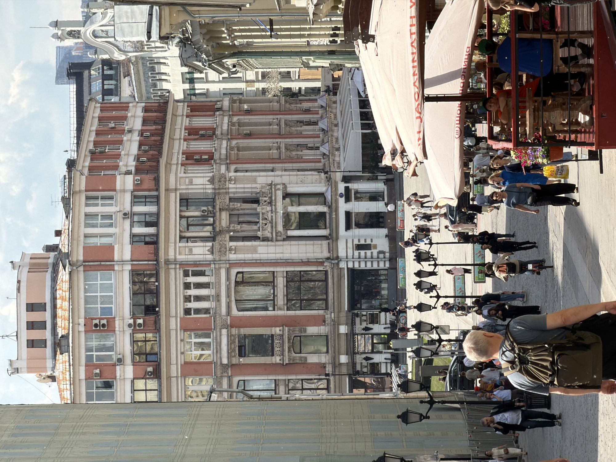 This image captures a vibrant, sunny daytime scene on a bustling street in Moscow, Russia. The perspective is slightly elevated, looking down the street.
On the left, a multi-story building stands prominently, featuring a distinctive architectural style with sections of reddish-brown brick alternating with ornate white or cream-colored facades. The building is adorned with numerous windows and a series of air conditioning units visible on its exterior. Further back, more buildings with decorative rooftops, some featuring domes, are visible against a partly cloudy sky.
To the right, an active outdoor cafe or restaurant extends into the street, with patrons seated under large, light-colored awnings. The word "JAGANNATH" is clearly visible in large letters on one awning. Another awning displays text in Cyrillic, likely "ШОКОЛАДНИЦА" (Shokoladnitsa), suggesting a chocolate cafe. People are seen dining and socializing at the cafe, which is decorated with flower planters.
The street itself is a pedestrian-heavy area, lined with numerous black, multi-armed lampposts stretching into the distance. Many people are walking along the street; in the foreground, a man with white hair, wearing a grey shirt and a dark green backpack, is walking away from the camera. Other individuals, including someone on a scooter, are dispersed throughout the scene. A reflective surface in the lower foreground mirrors portions of the street and adjacent structures. A yellow bag carried by a person in the mid-ground shows Cyrillic text, possibly "МОРОЖЕНОЕ" (Ice cream). The bright sunlight and visible shadows indicate it is daytime, and the weather appears clear and pleasant.