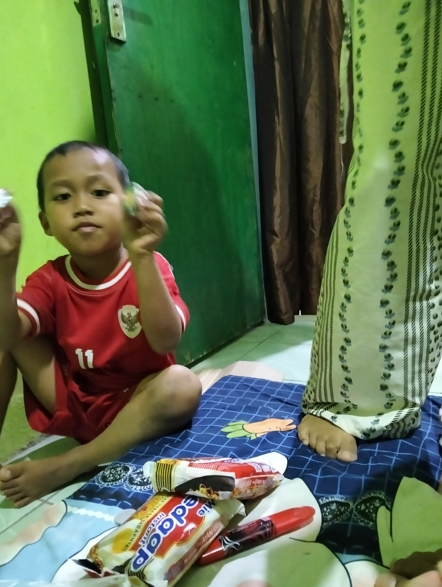 A young boy in a red shirt with white trim and the number "11" on the front is sitting cross-legged on a blue and white checkered blanket. He is holding two small, translucent objects in his hands. To his right, a person's legs are visible, wearing a light green patterned garment with vertical stripes and floral accents. Several packaged food items, including what appears to be instant noodles and a red tube, are scattered on the blanket in front of the boy. The background features a bright green wall on the left and a dark brown curtain on the right, with a glimpse of a door handle on the green wall. The scene appears to be indoors.