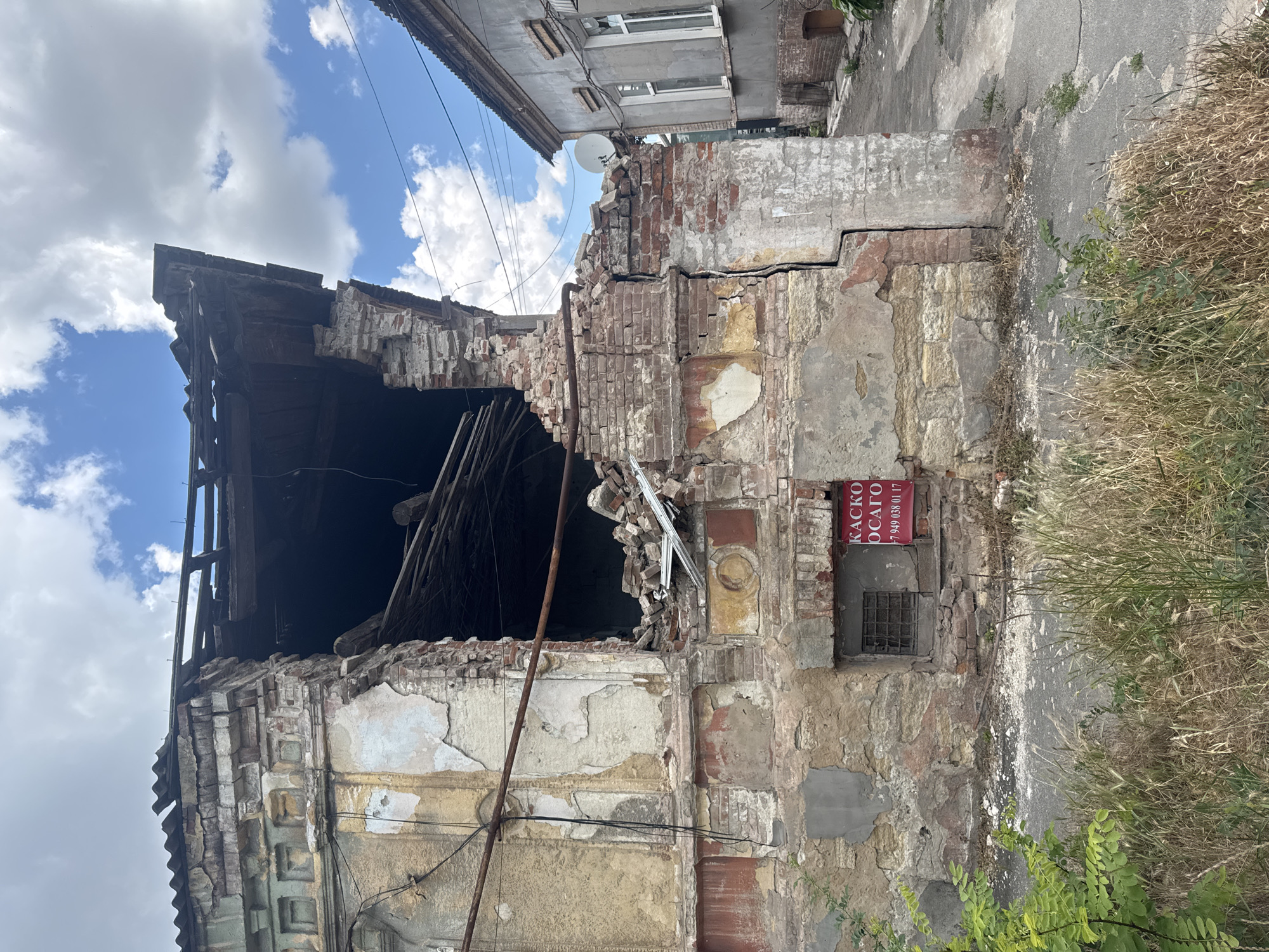 The image shows a heavily damaged building in Mariupol, Ukraine. The building is made of brick and plaster, with a collapsed roof and a large hole in the side. There is debris scattered around the building, including bricks and wooden beams. In front of the building is a small window with a metal grate and a red sign with Ukrainian text and a phone number. The sky is blue with white clouds, and there is some vegetation in the foreground. To the right of the damaged building is another building with a satellite dish on its roof. The overall scene suggests a recent event of destruction.