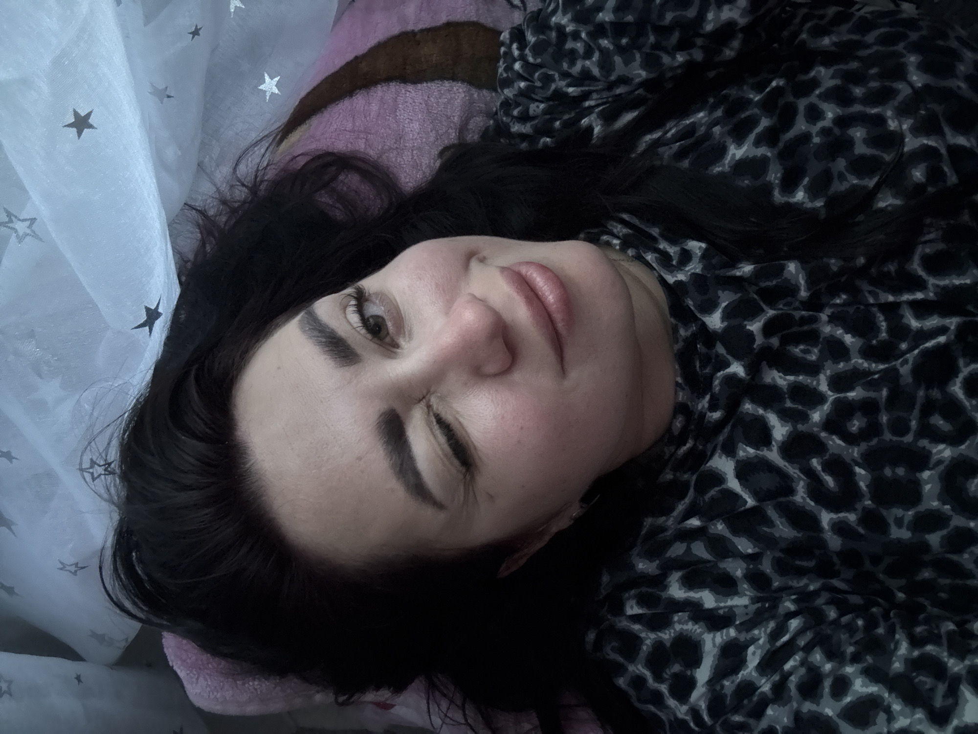 A woman with dark hair and makeup is lying down, winking at the camera. She is wearing a black and grey leopard print top. The background consists of a pink pillow and a white fabric with silver stars. The scene appears to be indoors.