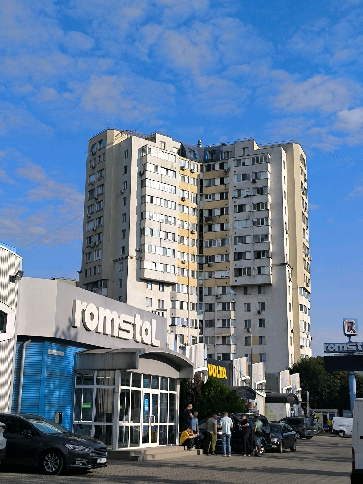 The image shows a tall, multi-story residential building in Chișinău, Moldova, under a bright blue sky with scattered clouds. In the foreground, there is a commercial building with a prominent "romstal" sign. Several people are standing around near the entrance of the "romstal" building and a few cars are parked in front. To the right, a "VOLTA" sign is visible on another structure. Further to the right, a gas station sign with a red "R" logo is also present. The residential building is a typical example of Soviet-era architecture, characterized by its repetitive window patterns and concrete facade, with some balconies and air conditioning units visible. The overall scene depicts a busy commercial and residential area on a clear day.