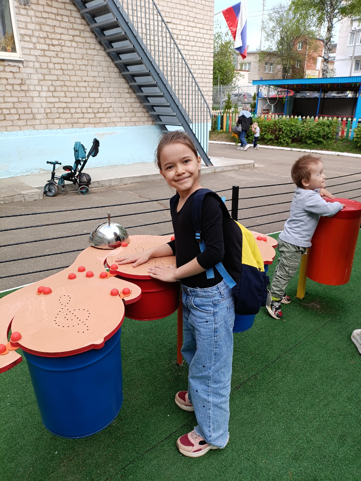 Observed is an outdoor scene in what appears to be a play area, likely situated in Chaikovsky, Russia, based on the visible Russian flag.
A young girl stands to the left of the frame, wearing a black long-sleeve shirt, light blue denim jeans, and sneakers. She has a backpack slung over her shoulder and is smiling towards the camera. Her hands are resting on a large, multi-colored play structure that resembles a drum set. The structure consists of a blue cylindrical base, a red cylindrical base, and a brown top with red knobs. A metal dome-shaped instrument sits on top of the structure.
To the right, a toddler leans against a red cylindrical structure supported by yellow legs. He wears a grey sweatshirt, khaki pants, and sneakers, gazing to his right.
In the background, a brick building is visible with a gray metal staircase attached to its side. To the left of the building, a blue tricycle sits on a paved area. Further in the background, a fence with colorful pencil-shaped posts separates the play area from an adjacent area with more buildings, greenery, and people. A woman and child are walking in the background. The ground is covered in green artificial turf.
