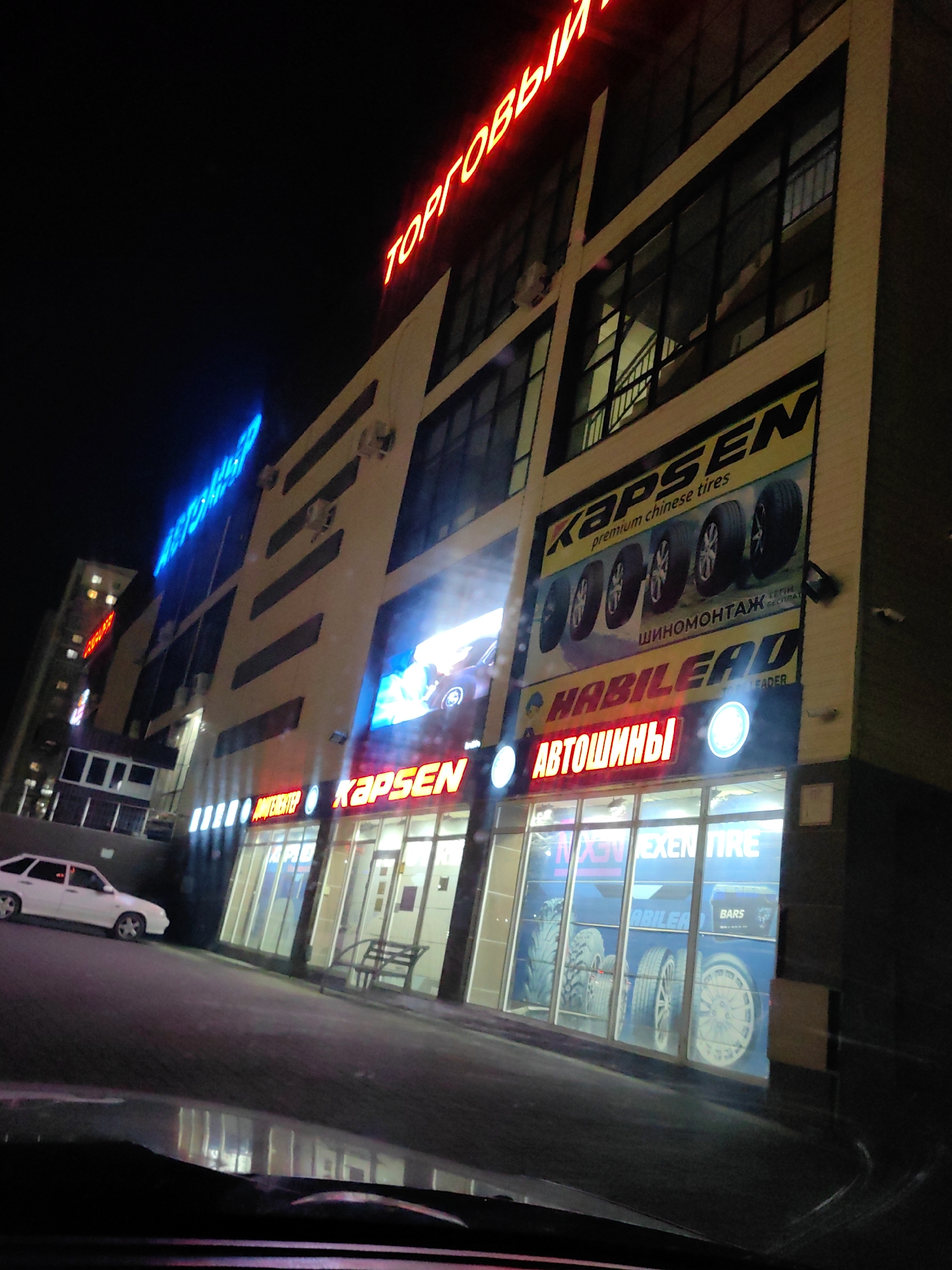 The image is a nighttime street-level shot of a commercial building in Shymkent, Kazakhstan, likely a tire shop. The building has multiple stories with large windows and illuminated signage.
Prominently displayed are several signs. At the top, in red neon letters, is signage that reads "ТОРГОВЫЙ" (Torgovy), meaning "Trade" or "Commercial." Below this, a large banner advertises "KAPSEN premium chinese tires," featuring images of car tires. Further down, another sign in red letters reads "АВТОШИНЫ" (Avtoshiny), meaning "Autotires."
Other visible signage includes blue neon lettering on the upper floors and advertisements for "HABILEAD" tires on a lower banner. The storefronts feature large glass windows displaying tire advertisements and possibly tire products. A white car is parked in front of the building on the asphalt road. The environment appears to be a typical urban street at night, with artificial lighting from the building and streetlights. The overall scene suggests a commercial district or a road with businesses.