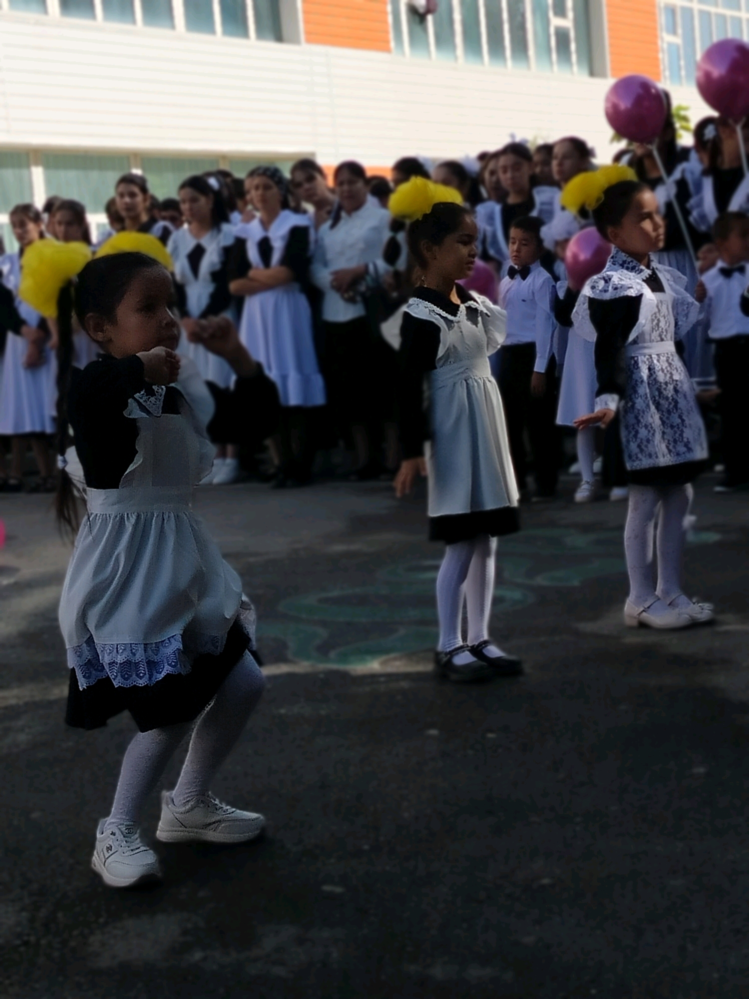 In the city of Kölkent, Kazakhstan, a group of schoolchildren, predominantly girls wearing traditional school uniforms consisting of dark dresses with white pinafores and white tights, are gathered outdoors. In the foreground, a young girl with dark hair, seen in profile, is in motion, her left arm raised. She is wearing a dark dress, a white pinafore with blue lace trim, white tights, and white sneakers. To her right, another girl in a similar uniform is also in motion, her right hand extended forward. She wears a dark dress, a white lace pinafore, white patterned tights, and white shoes. Behind them, a crowd of schoolchildren and adults are visible. Some children are holding balloons, including pink and purple ones. The background features a modern building with white and orange sections and windows. The ground is paved with dark asphalt. The scene appears to be an outdoor school event, possibly a celebration or assembly.