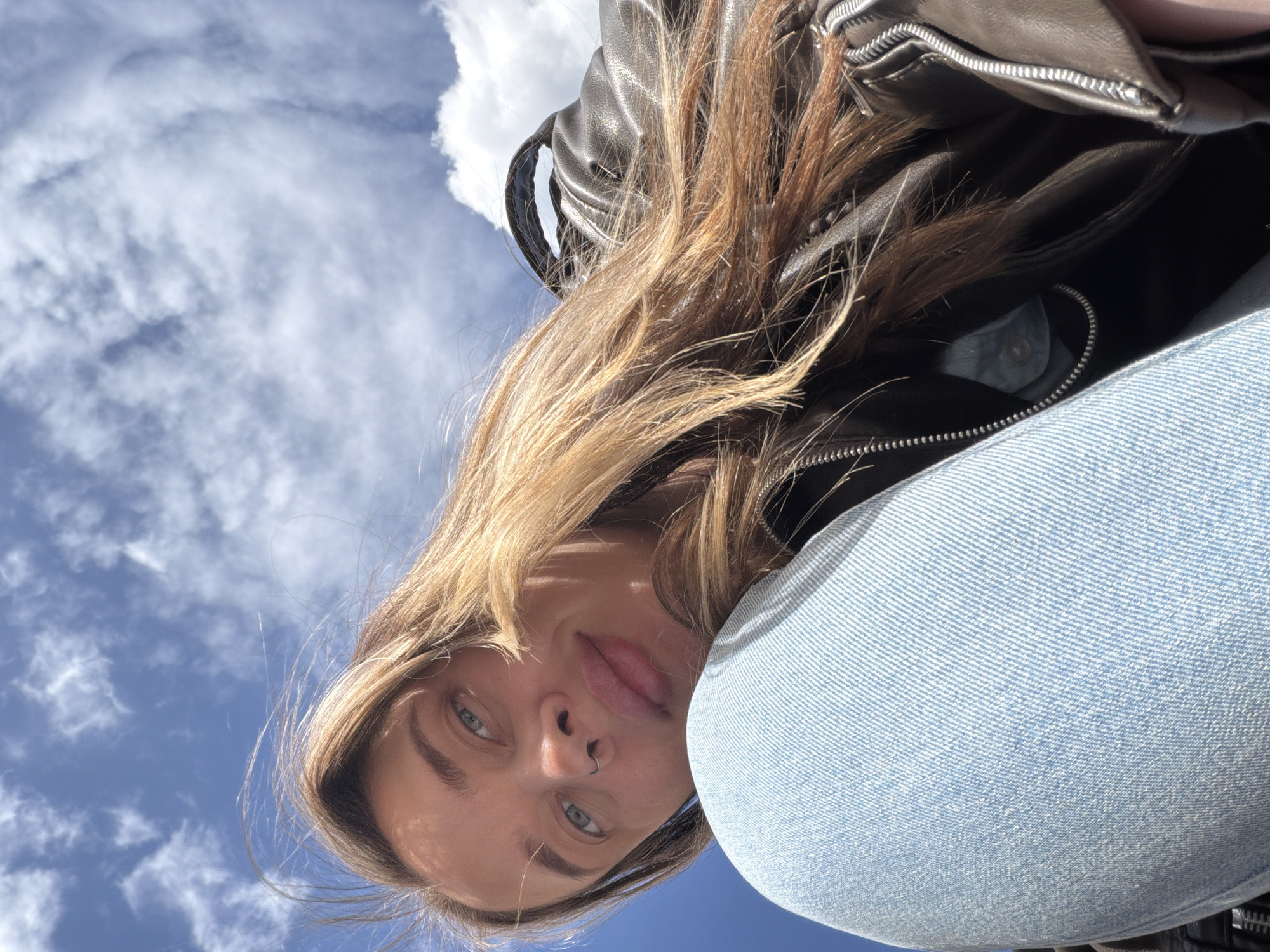 A woman with blonde hair, wearing a black leather jacket and light blue jeans, is looking down at the camera against a backdrop of a blue sky with white clouds. She has a nose ring. The location cannot be determined as Moscow, Russia, from the provided image.