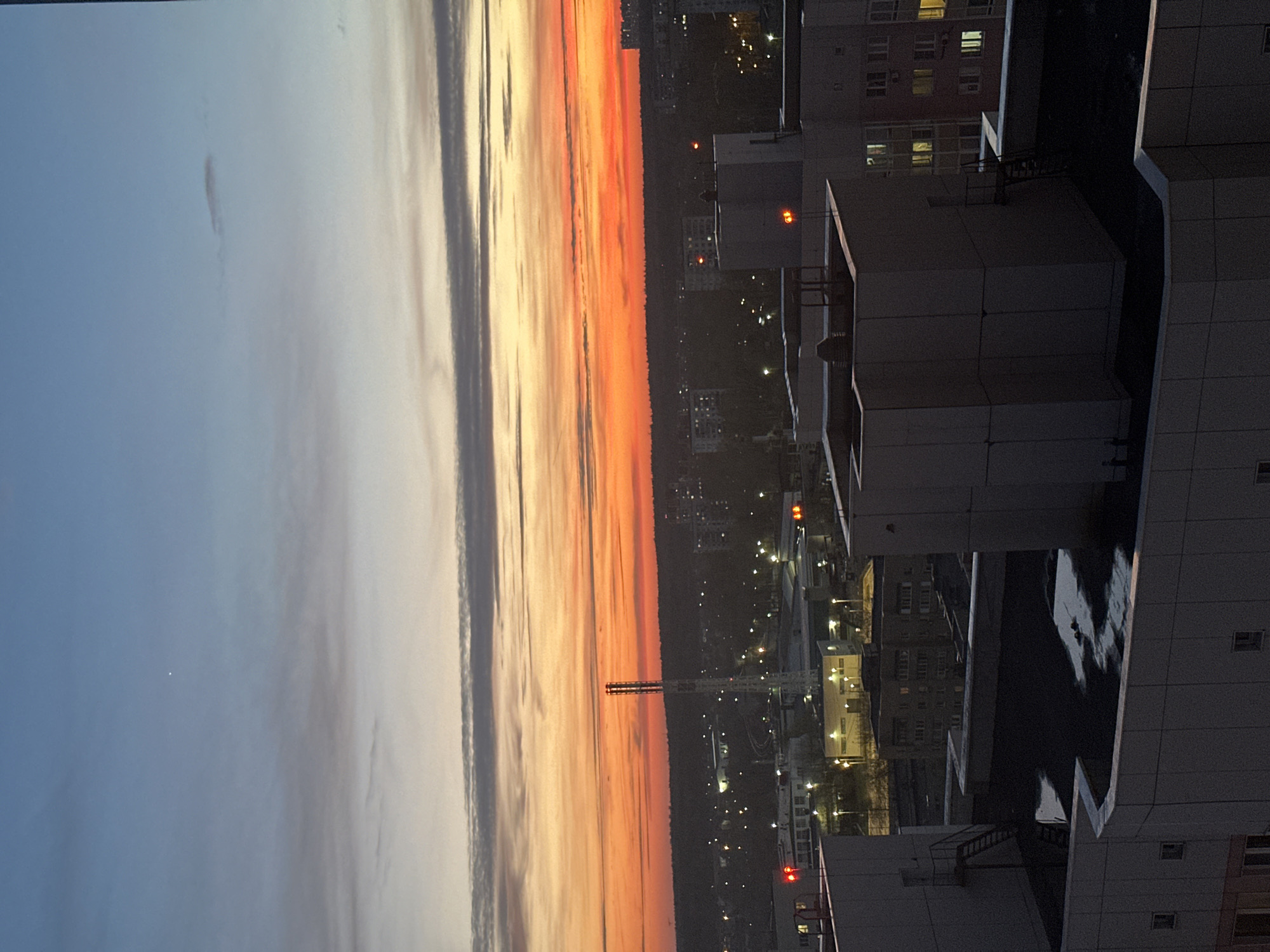 A cityscape in Moscow, Russia at sunset, viewed from an elevated vantage point. The sky transitions from blue on the left to orange and yellow toward the horizon. Buildings line the lower right portion of the image, showing lit windows and structures with a rooftop. A tower is seen in the distance. The city appears illuminated by artificial lights scattered among the buildings.