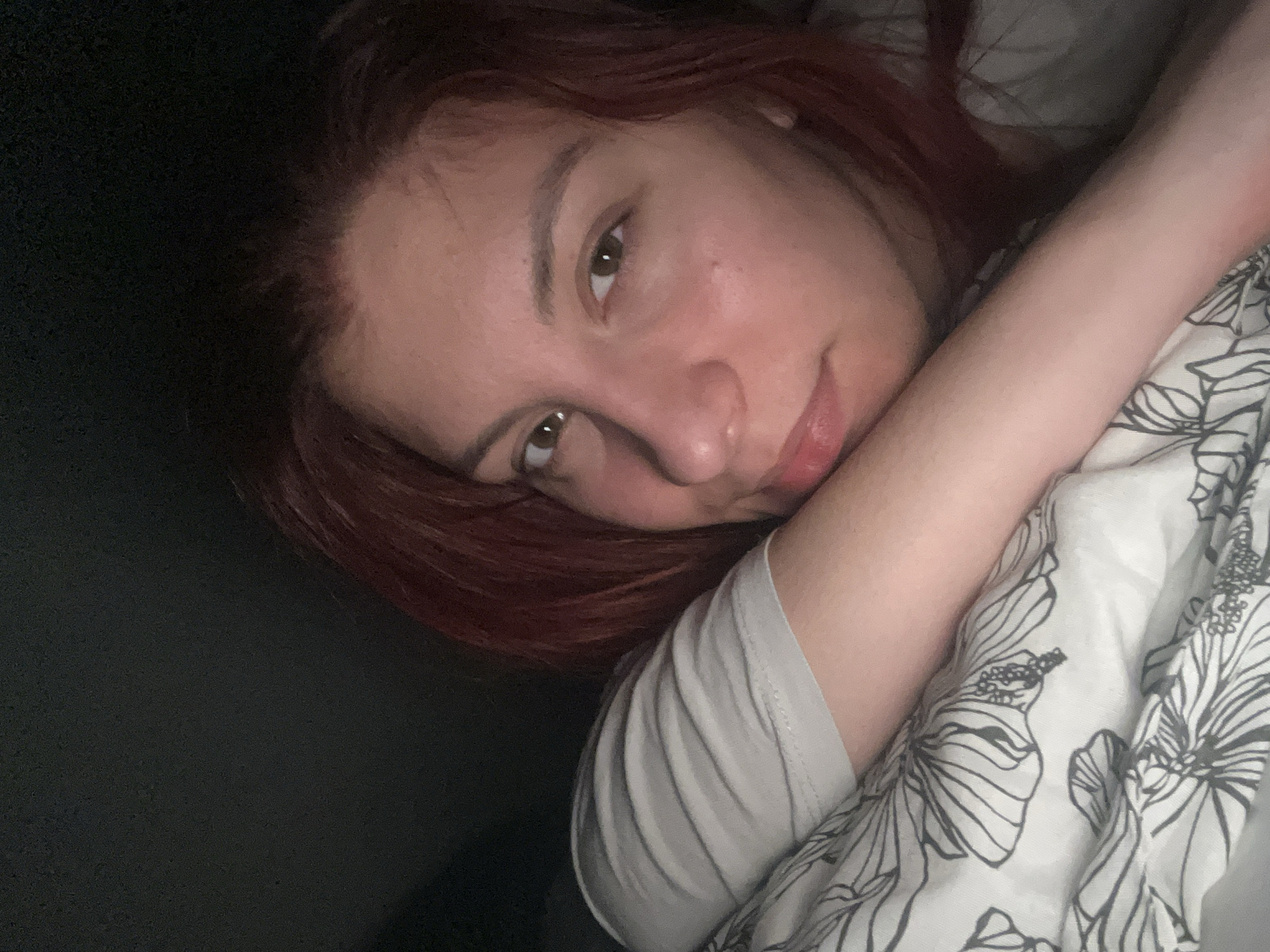 A person with reddish-brown hair is lying down, their head angled towards the camera. They have fair skin and brown eyes. Their right arm is bent and rests on a white pillow or bedding with a black floral pattern. The person is wearing a light grey t-shirt. The background is dark and indistinct. The location cannot be determined.