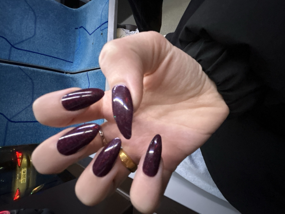 A close-up shows a person's hand with long, almond-shaped nails painted a deep burgundy color with subtle glitter flecks. The hand has a gold ring on one finger and a silver ring on another. The background includes a blue and gray surface, a glimpse of a car, and a dark fabric sleeve. The focus is on the hand and the glossy, jewel-toned nails.