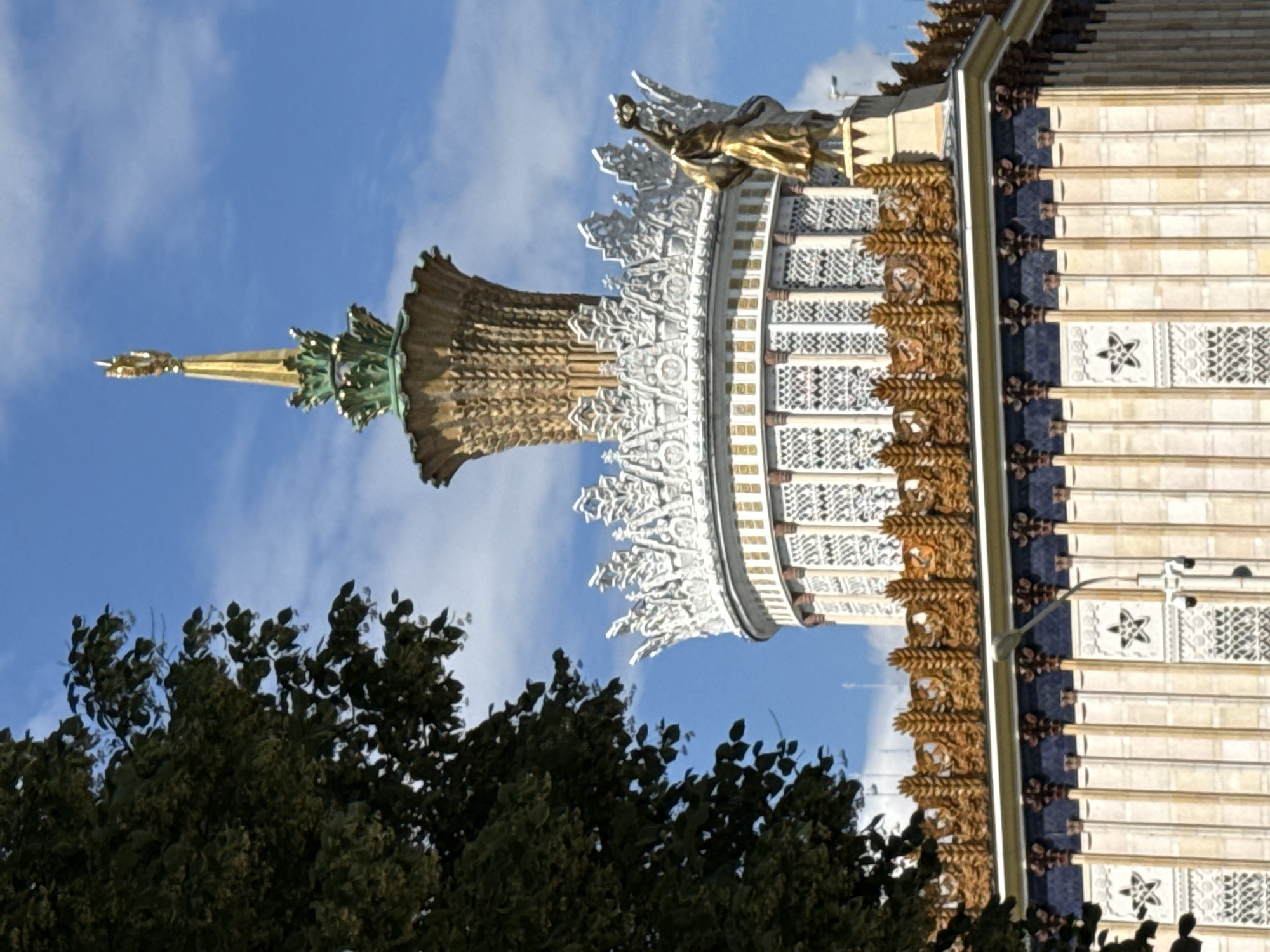 This is a close-up, rotated photograph of the upper section of a grand building in Moscow, Russia, against a bright blue sky with scattered clouds. The building's architecture is ornate and detailed, featuring a golden statue of a winged figure atop a decorative spire. Below this, intricate white scrollwork and metallic ornamentation adorn the building's facade. Further down, a textured section of the building displays a repeating pattern of stylized golden foliage. The main body of the building is composed of vertical cream-colored panels, each section separated by thin dark blue stripes and punctuated by decorative white starburst motifs. The overall impression is one of opulence and historical grandeur. The time of day appears to be midday, given the bright lighting and clear sky. The image is dominated by the architectural elements, with a dark green tree canopy visible in the foreground, partially obscuring the lower part of the building.